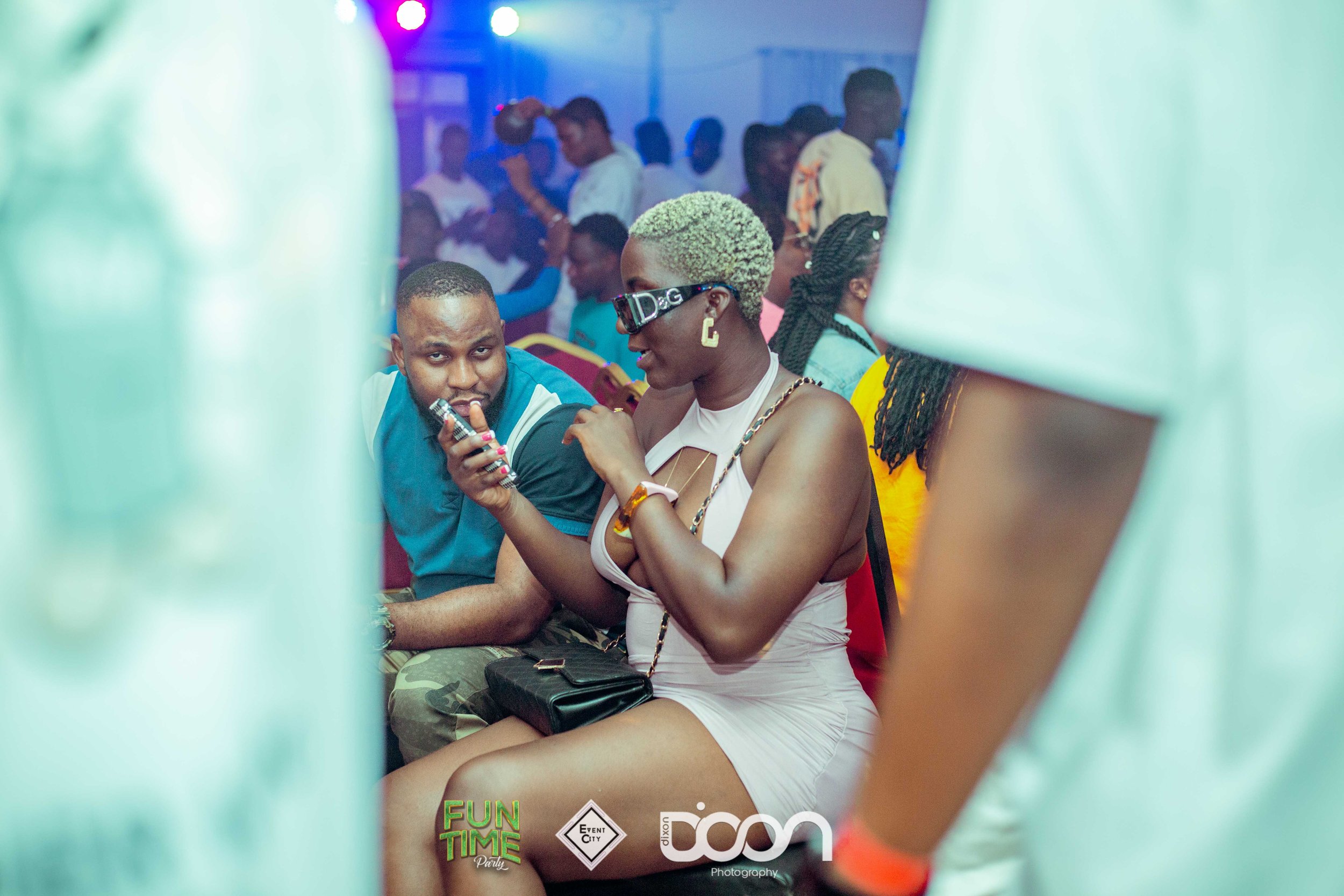 A woman wearing sunglasses, a pink dress, and gold accessories, sitting and showing her phone to a man at a party. The scene is lively with other people in the background.