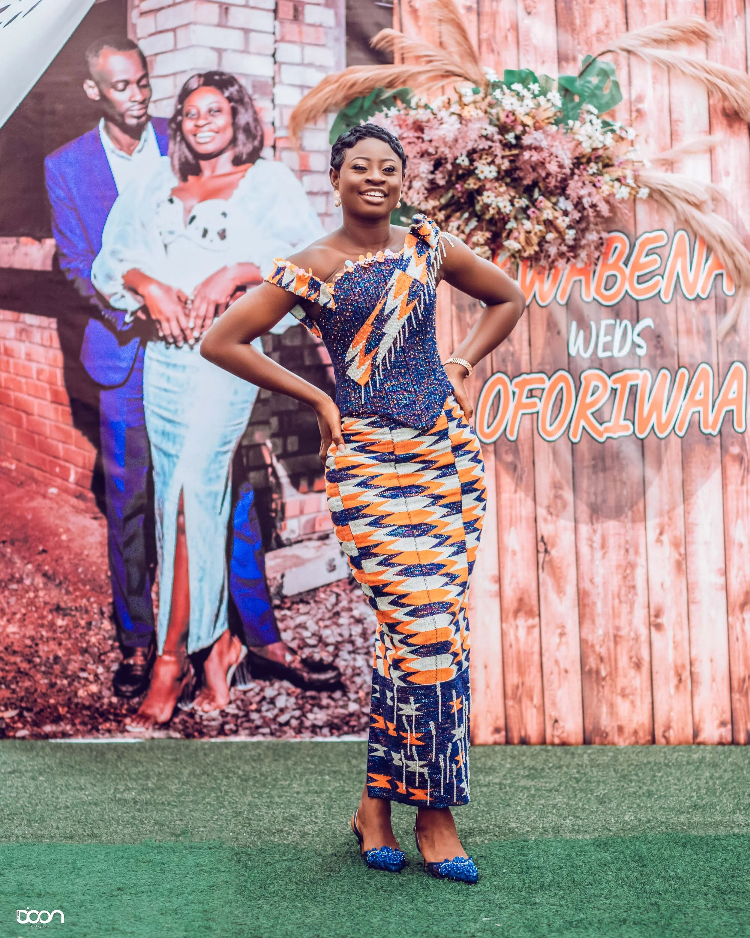 A woman in traditional African attire standing with her hands on her hips, smiling at an event with a floral and wooden backdrop that has text reading 'WUBENNA WEDS OFORIWA'.