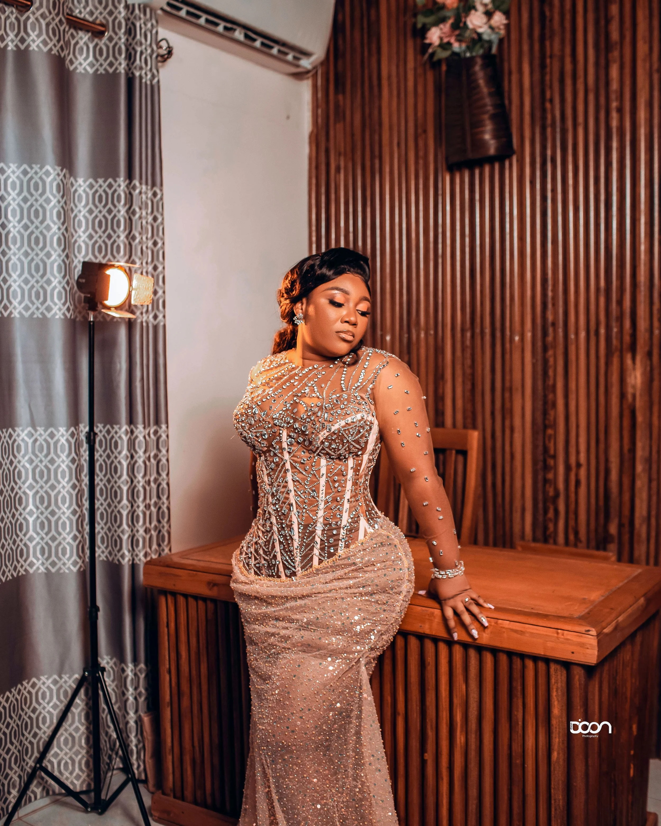 A woman in a glamorous, embellished gown posing indoors with wooden wall and curtains in the background.