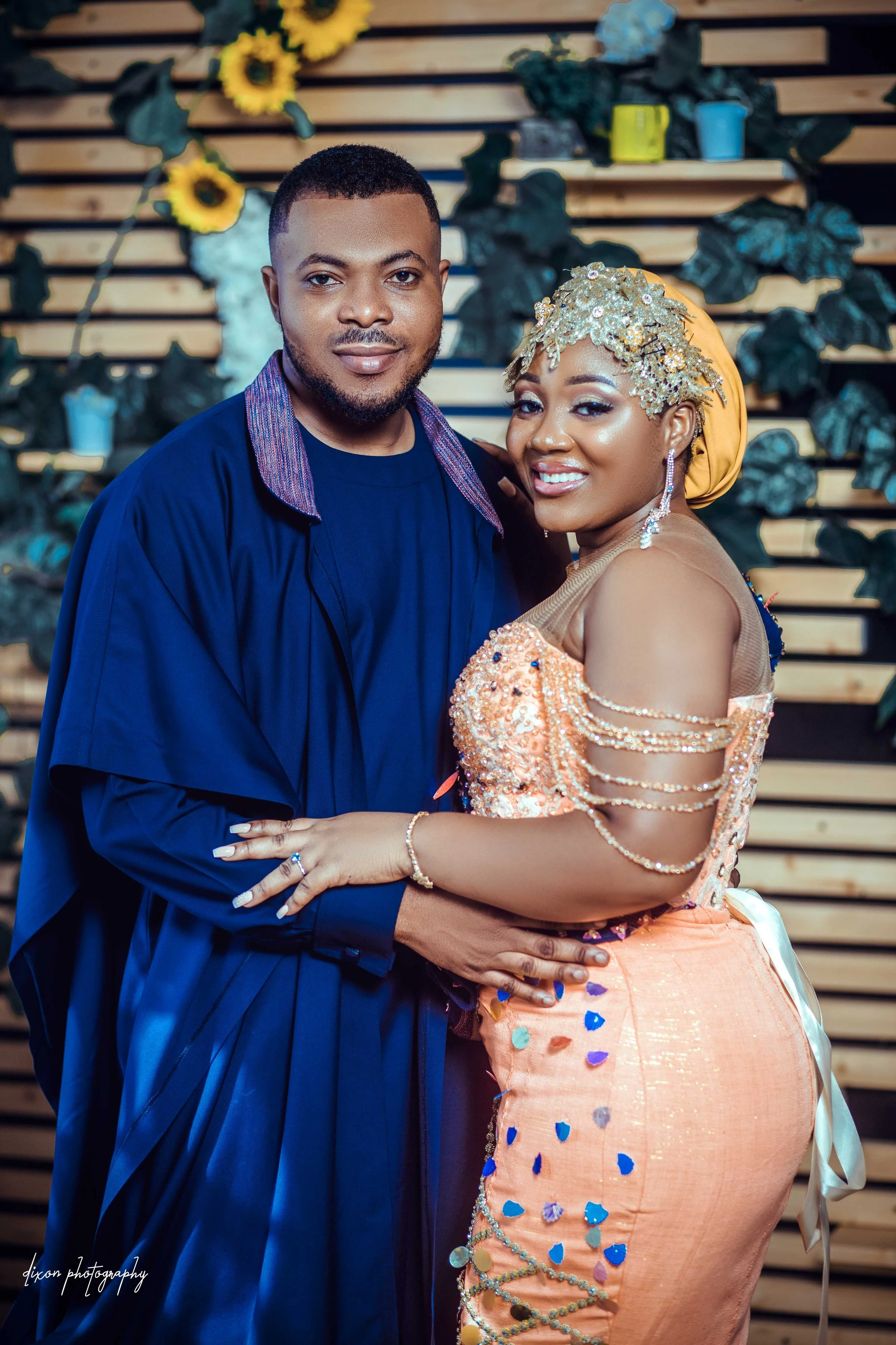 A smiling couple poses together at a celebration, with the man dressed in a dark blue outfit and the woman in an elaborate peach gown with bead embellishments and gold accessories, standing in front of a wooden backdrop adorned with sunflowers and gr