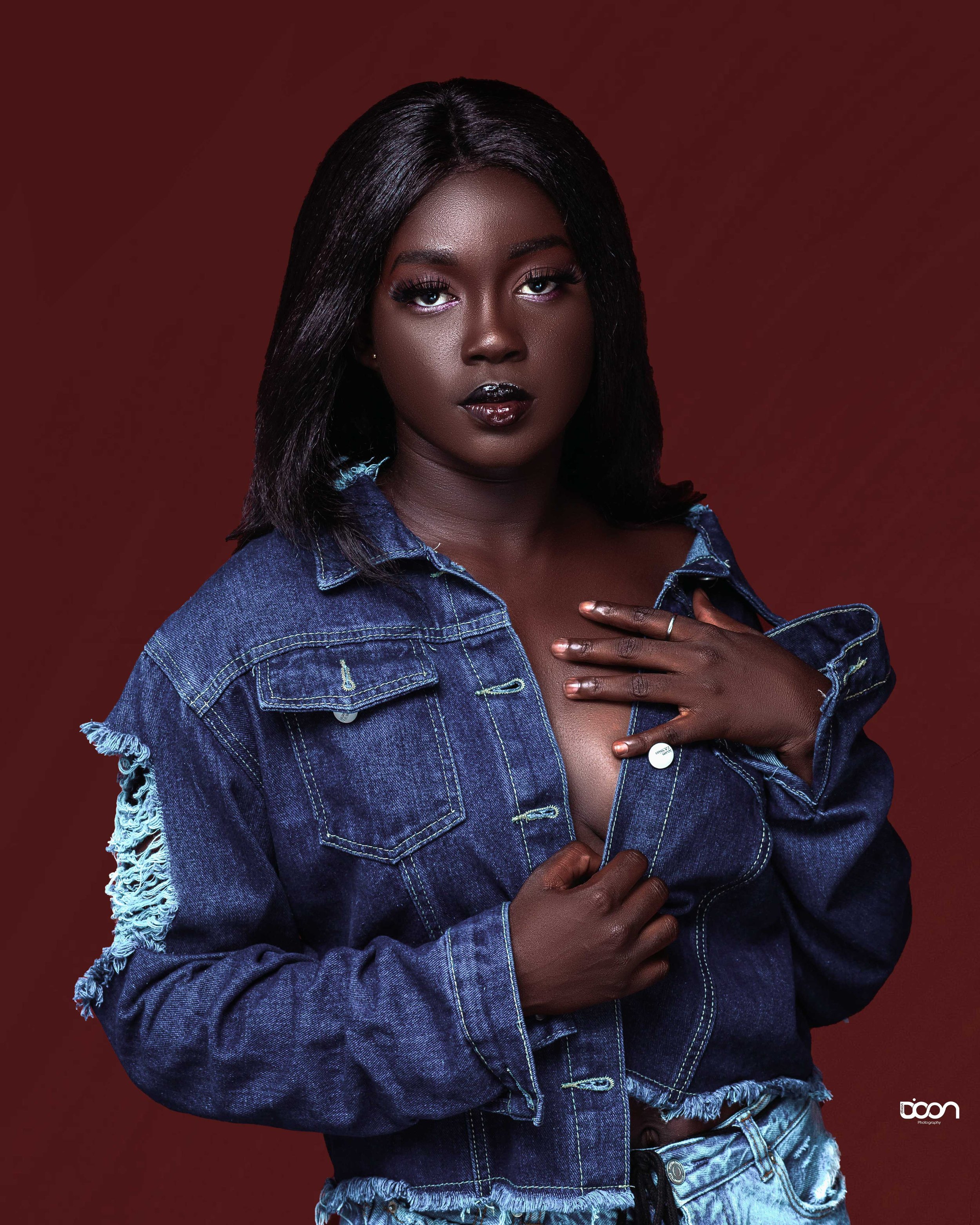 A woman with dark skin and straight black hair, wearing a distressed denim jacket and light-wash jeans, holding the jacket open to reveal her chest, against a solid dark red background.