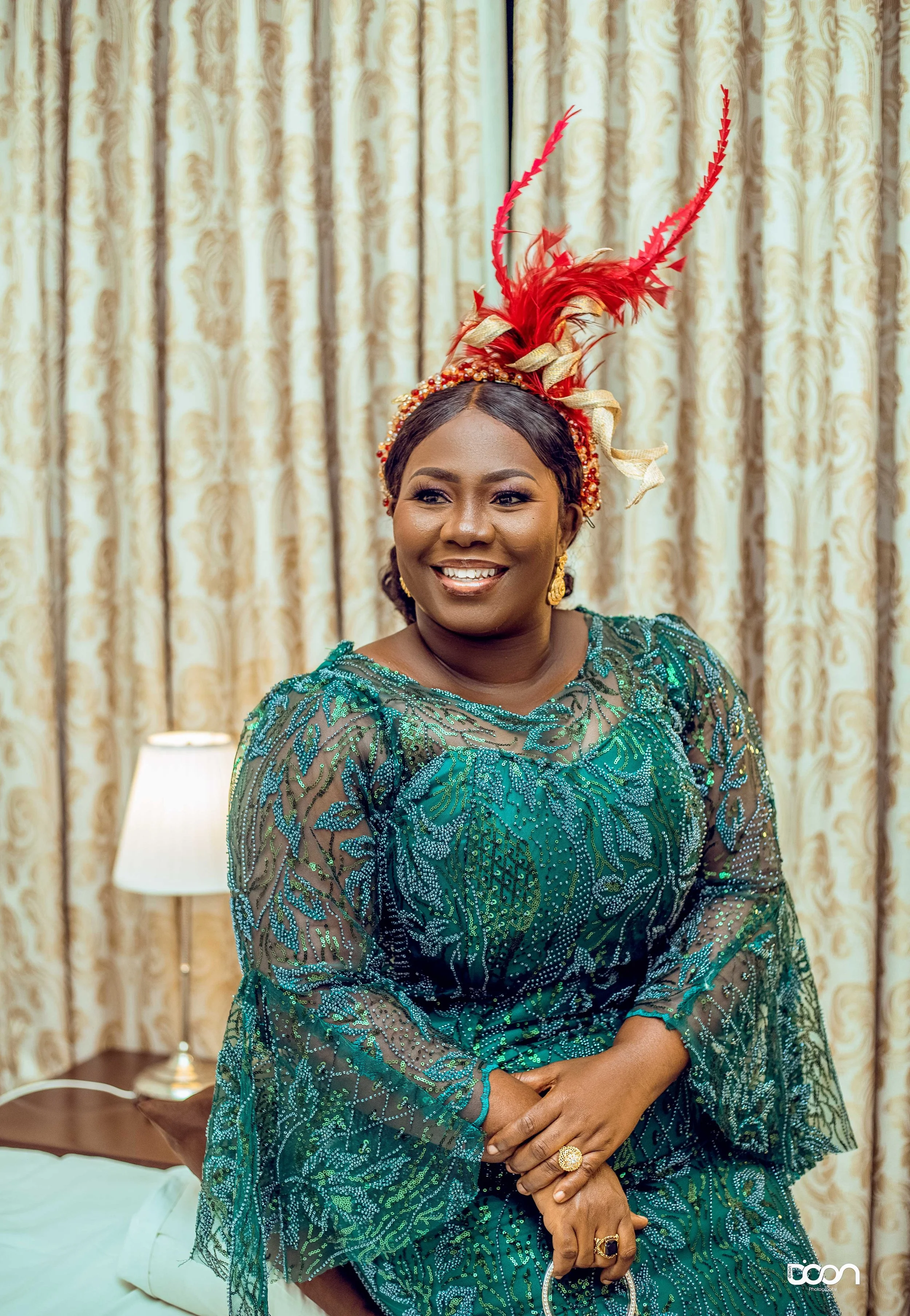 A woman in a green and blue patterned dress, smiling, wearing a decorative red feather headpiece and jewelry, standing in a room with cream-colored curtains and a lamp.
