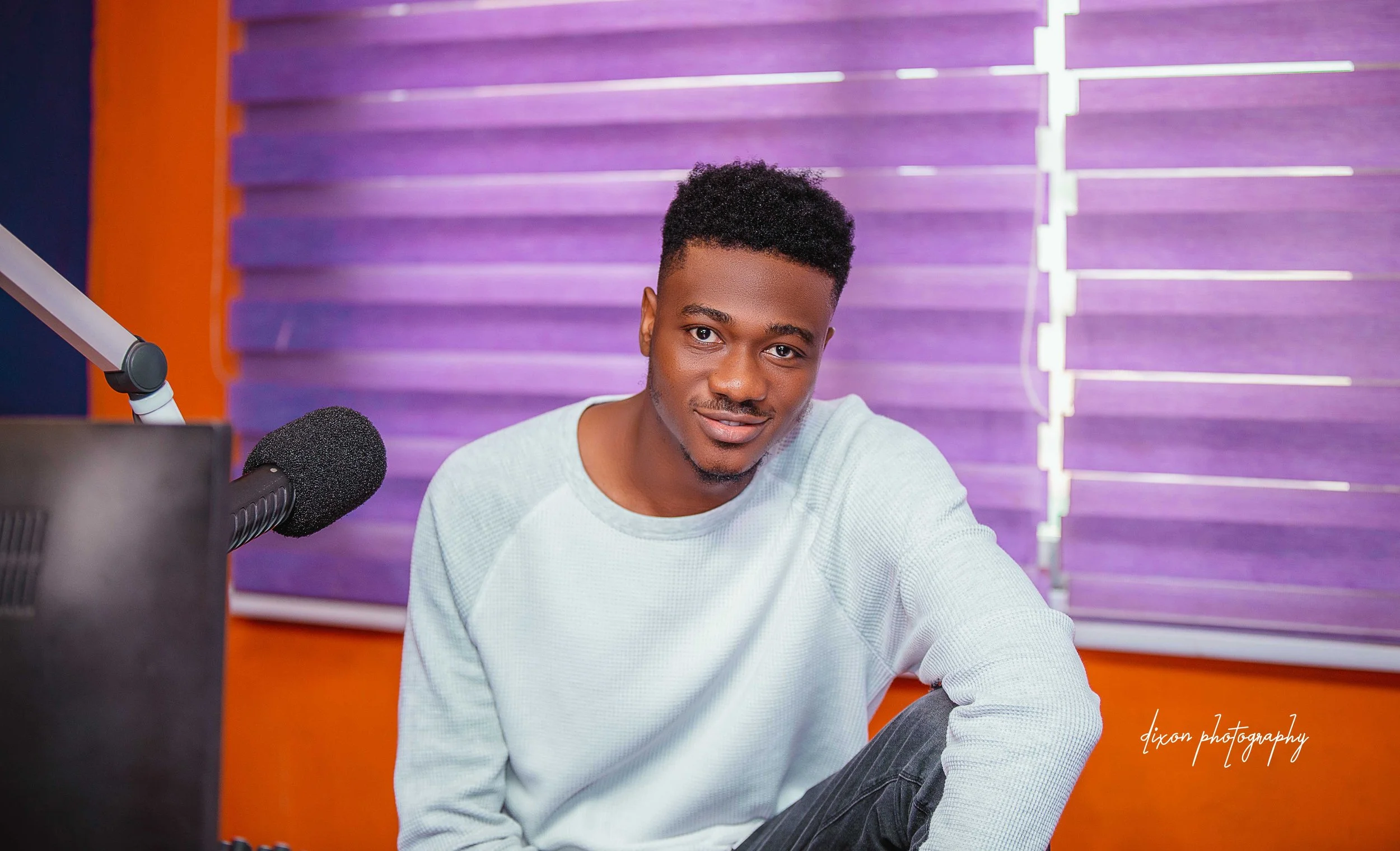 A young man with dark skin and short curly black hair sitting in a radio studio, smiling at the camera, with purple blinds in the background, a microphone on a stand to his left, and an orange wall behind him.