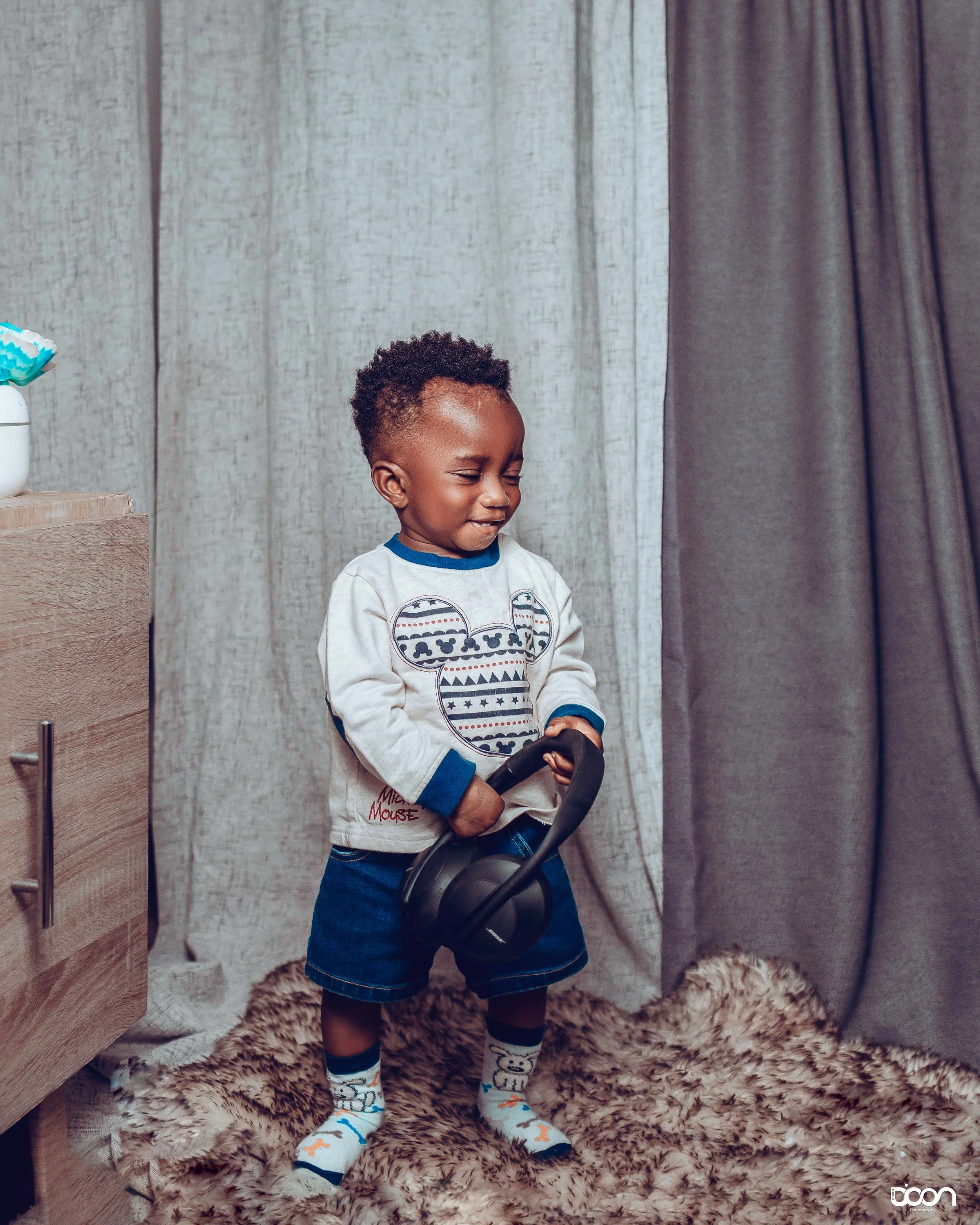 A young boy with curly hair, wearing a grey Mickey Mouse sweatshirt, denim shorts, and socks with bear designs, standing on a shaggy rug and holding a pair of black headphones, smiling with eyes closed.