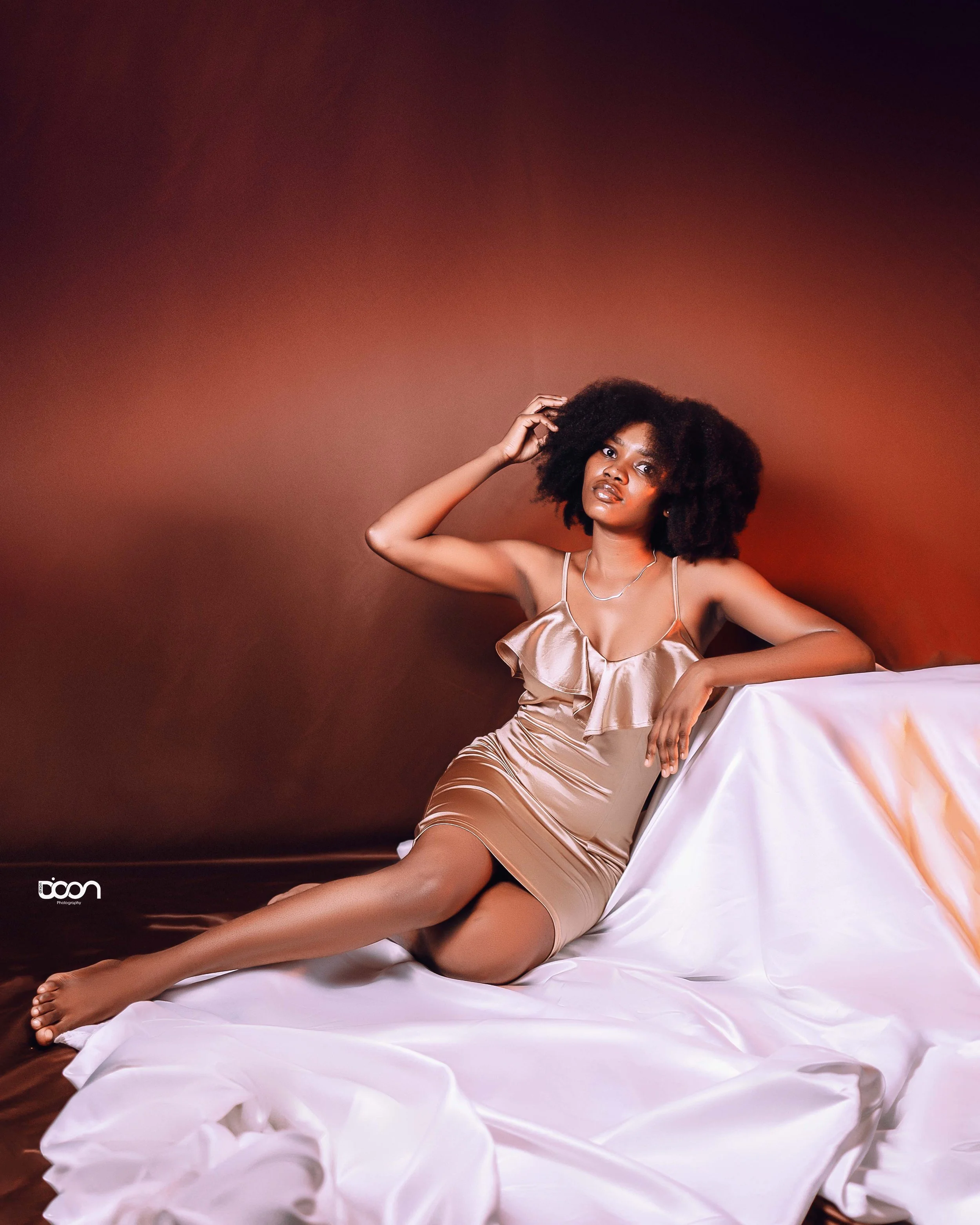 A woman with curly black hair sitting on a white fabric-covered surface against a brown background. She wears a beige satin dress, barefoot, and looks at the camera with one hand in her hair.