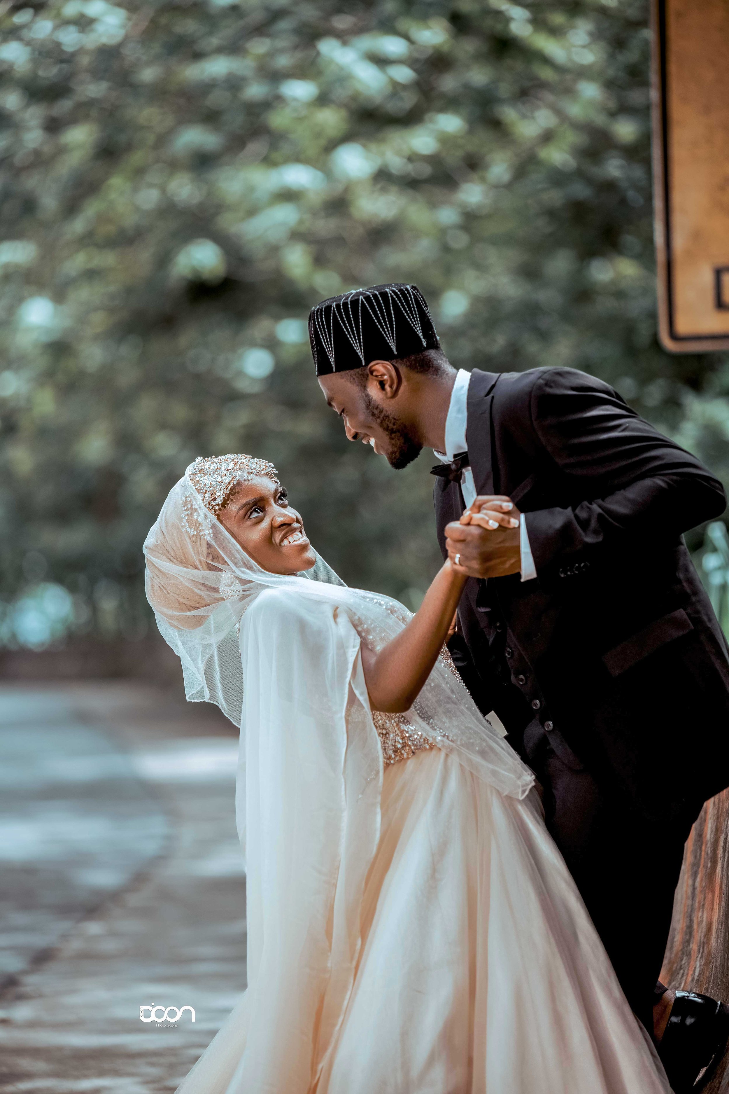 A joyful bride and groom sharing a dance outdoors, with the bride dressed in a white wedding gown and headscarf, and the groom in a black tuxedo, smiling at each other.