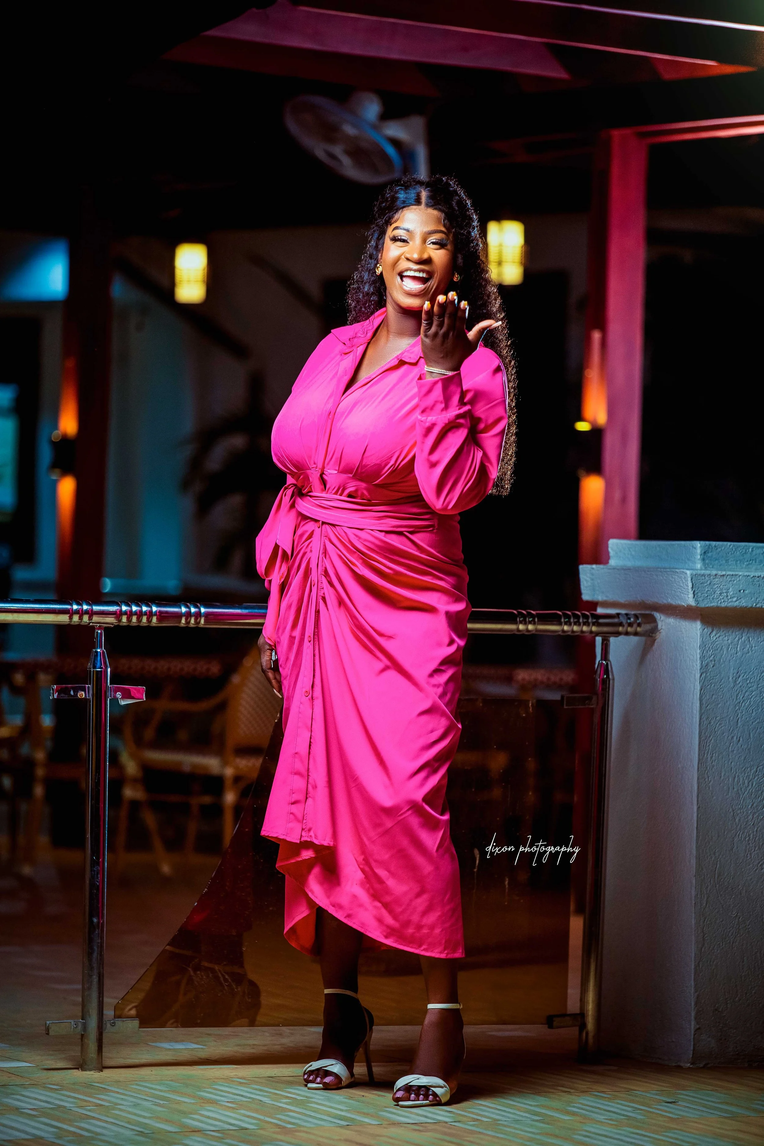 A woman in a bright pink dress stands on a wooden floor, smiling and blowing a kiss at night with warm indoor lighting.