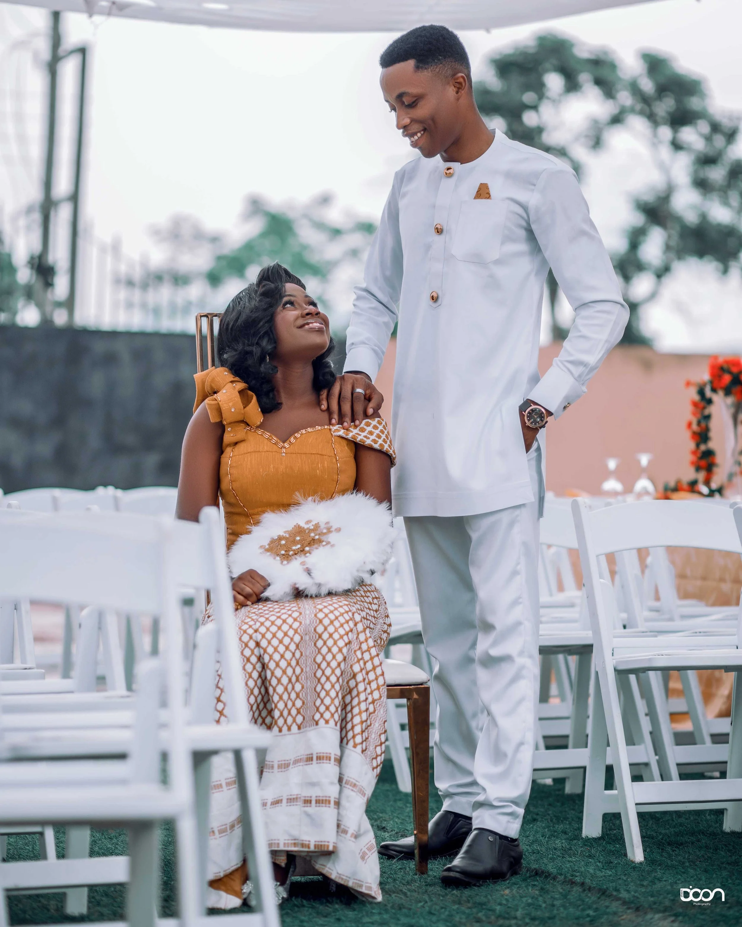 A woman in traditional attire sitting on a chair holding a fan, looking up at a man standing next to her with his hand on her shoulder, both smiling at each other, outdoors in a decorated venue.