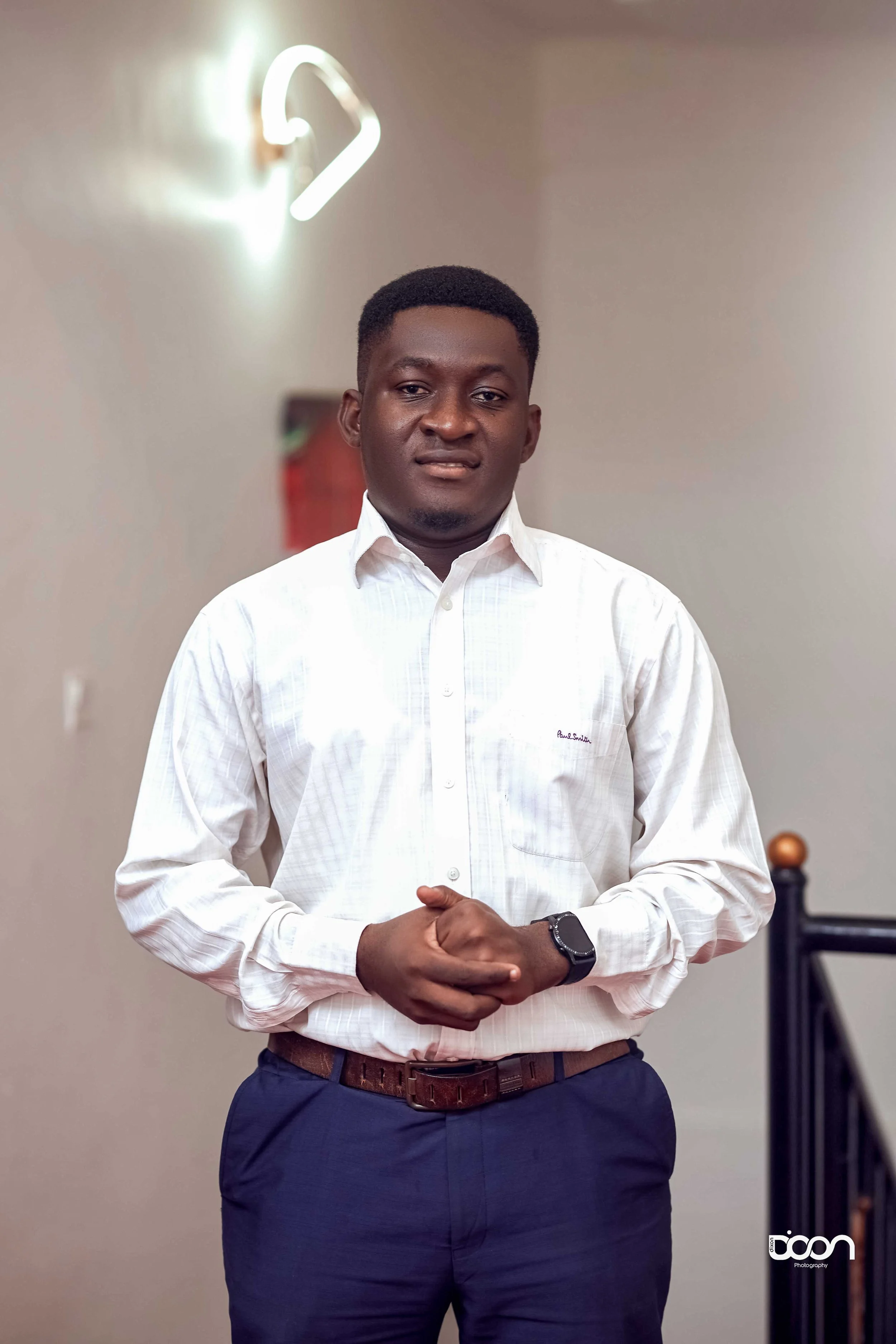 A man with dark skin and short black hair, wearing a white dress shirt and dark blue pants, standing indoors with hands clasped in front of him. The background includes a neon light and a neutral wall.