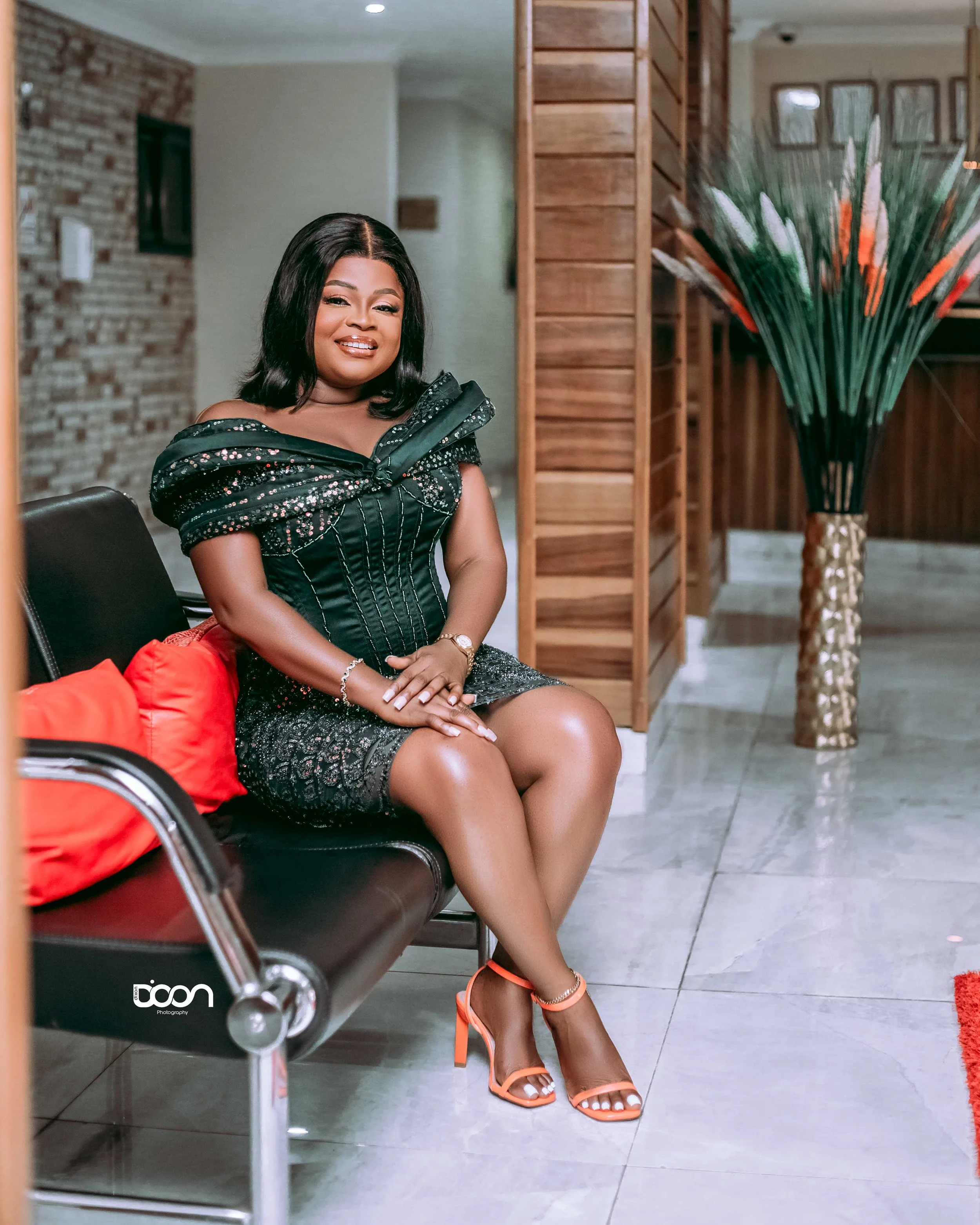 A woman in a black, shiny dress with sequins, sitting on a black bench with red pillows, wearing orange high heels, inside a room with wooden decor and a large vase with tall green and orange plants.
