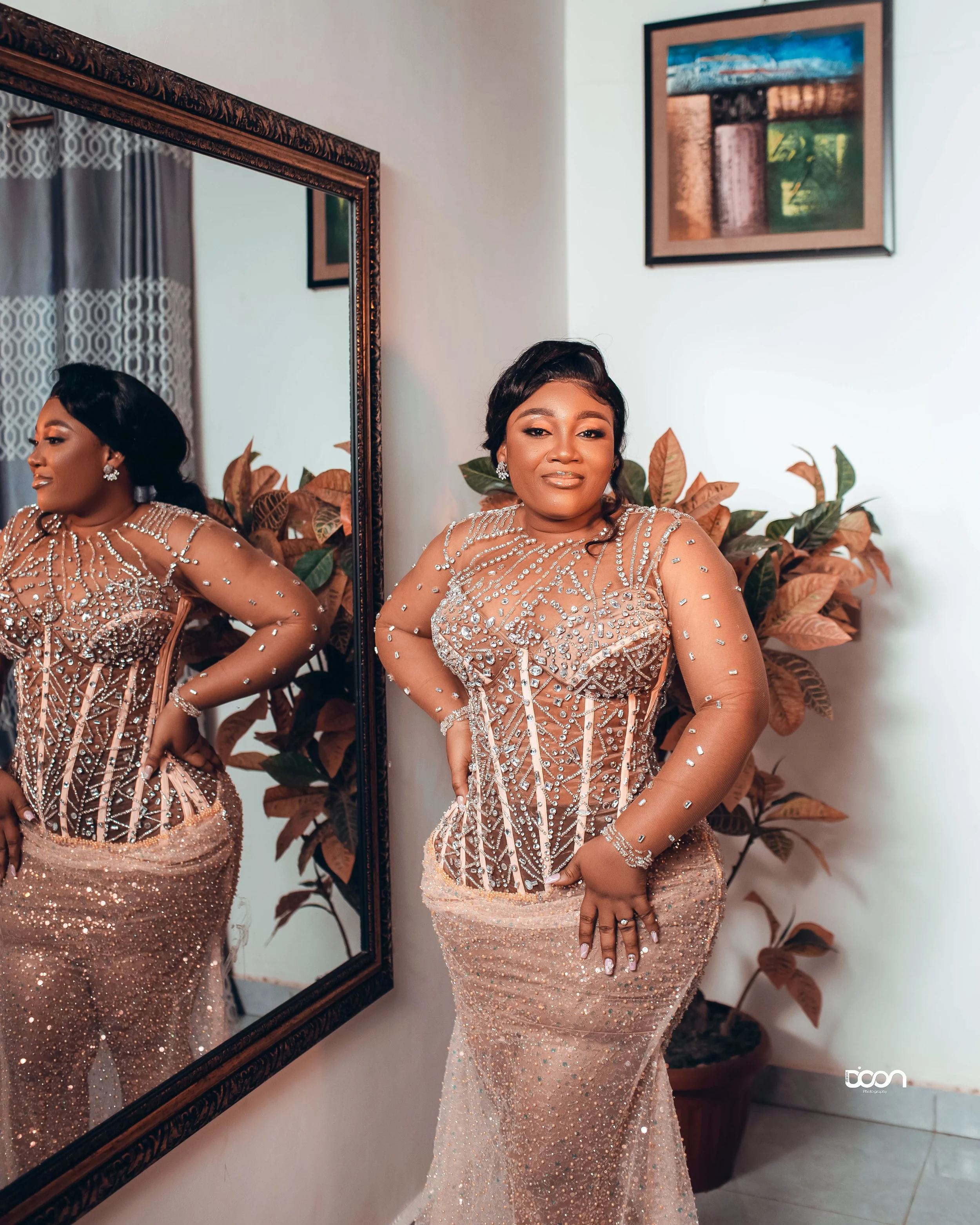 A woman in a glamorous, fitted, beaded dress standing next to a large mirror, with her reflection visible. She has dark hair styled elegantly and is wearing jewelry, standing in a room decorated with framed artwork and potted plants.