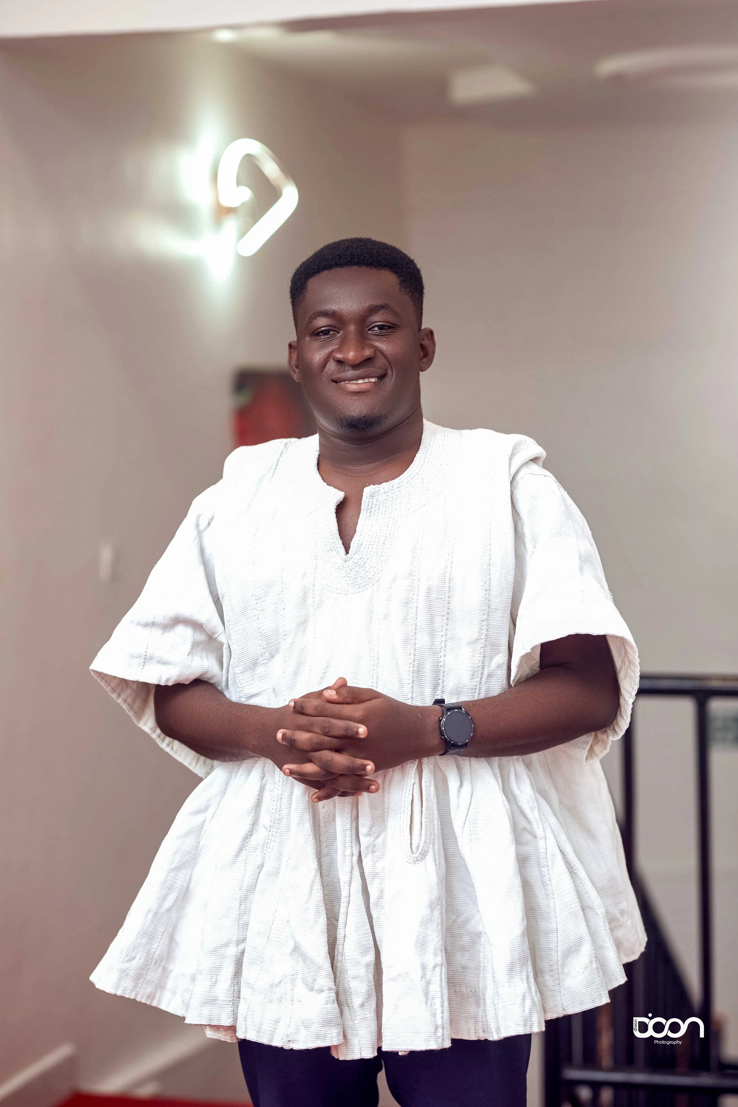 A man standing indoors wearing a white traditional African garment, smiling, with a black smartwatch on his wrist, and hands clasped in front of him.