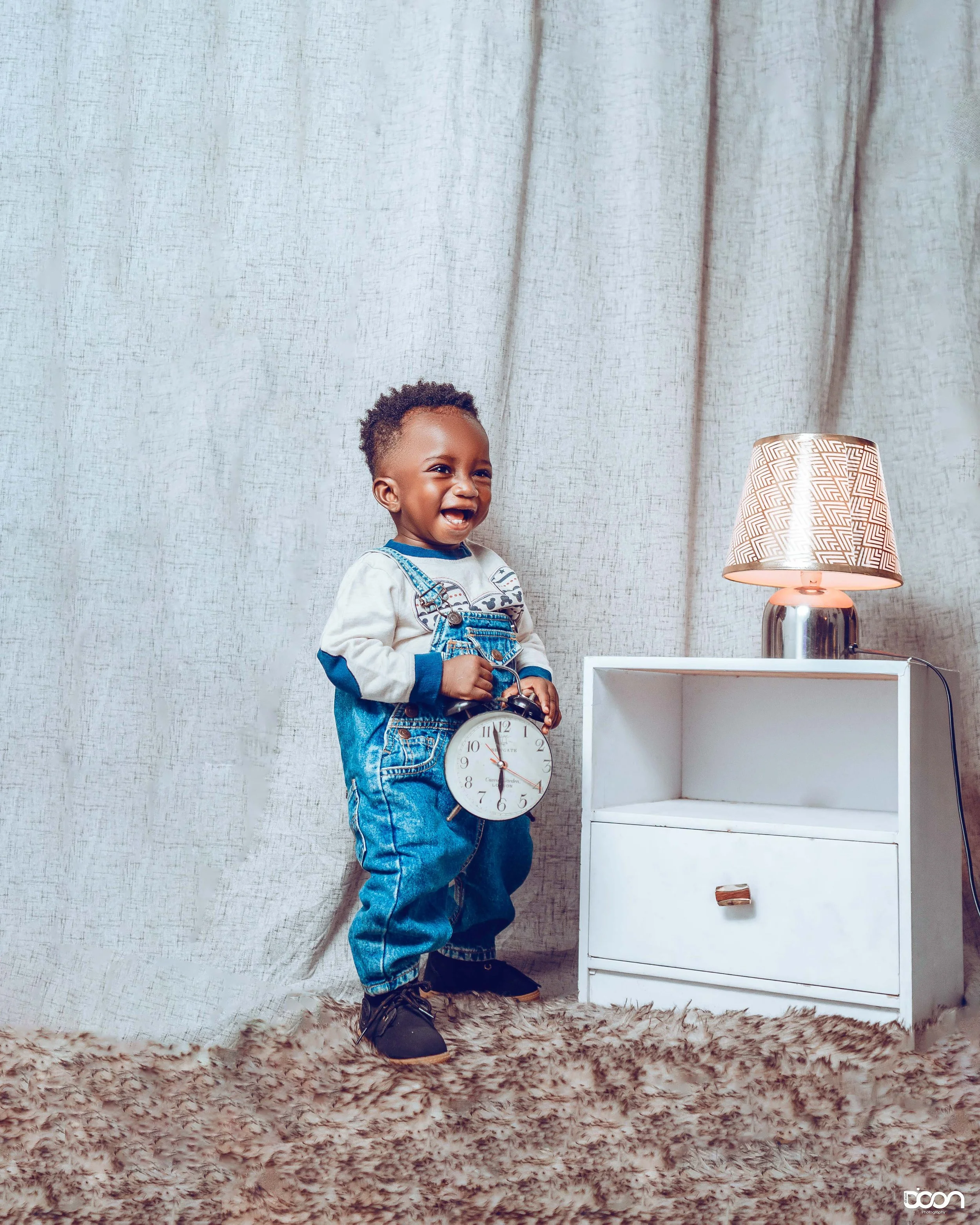 A young boy smiling and holding a clock, standing next to a white nightstand with a lamp on top, in front of a beige curtain and on a shaggy carpet.