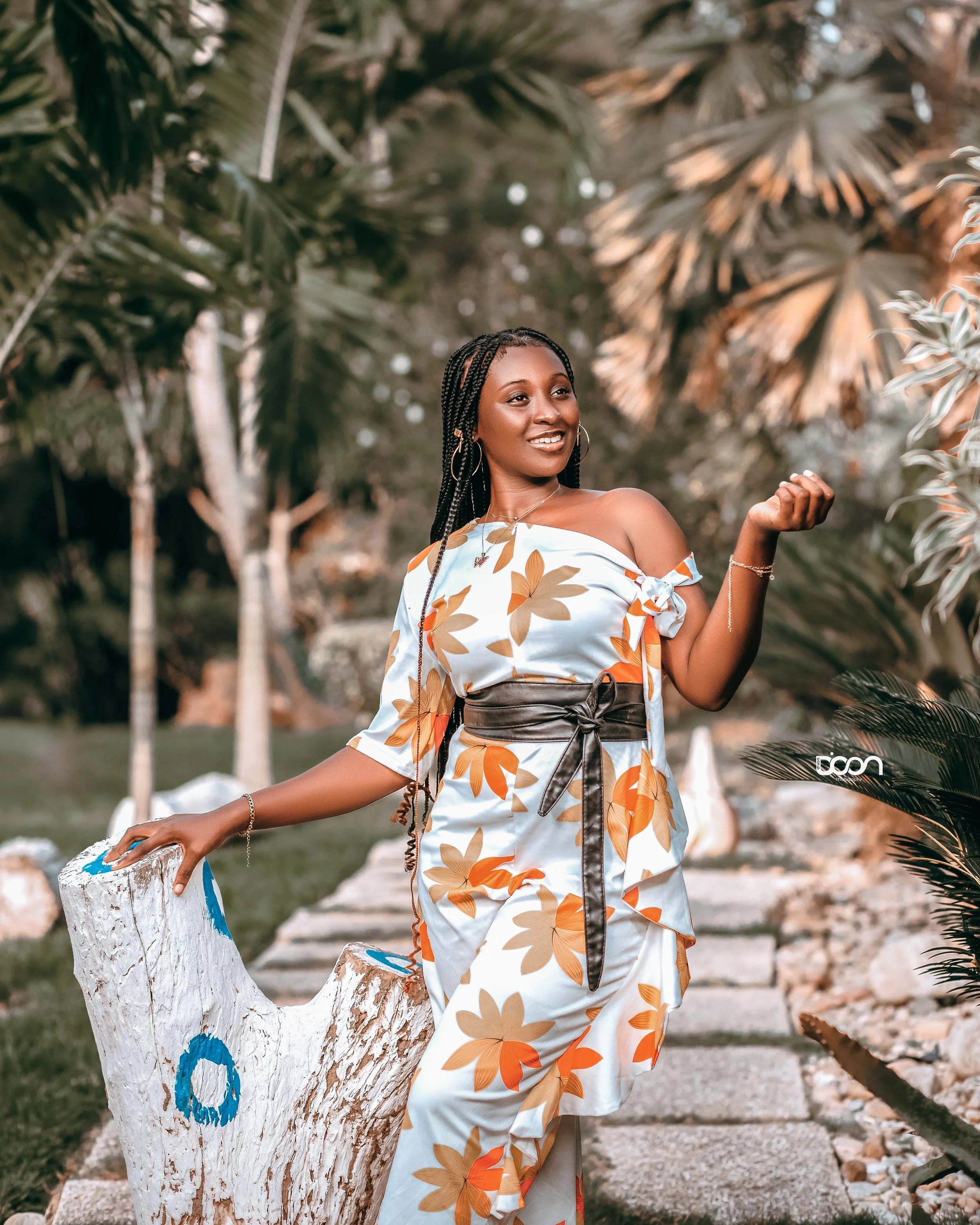 A woman in a floral dress with orange and beige leaves, standing outdoors in a garden with lush green trees, rocks, and a wooden log painted white with blue markings, smiling and posing.