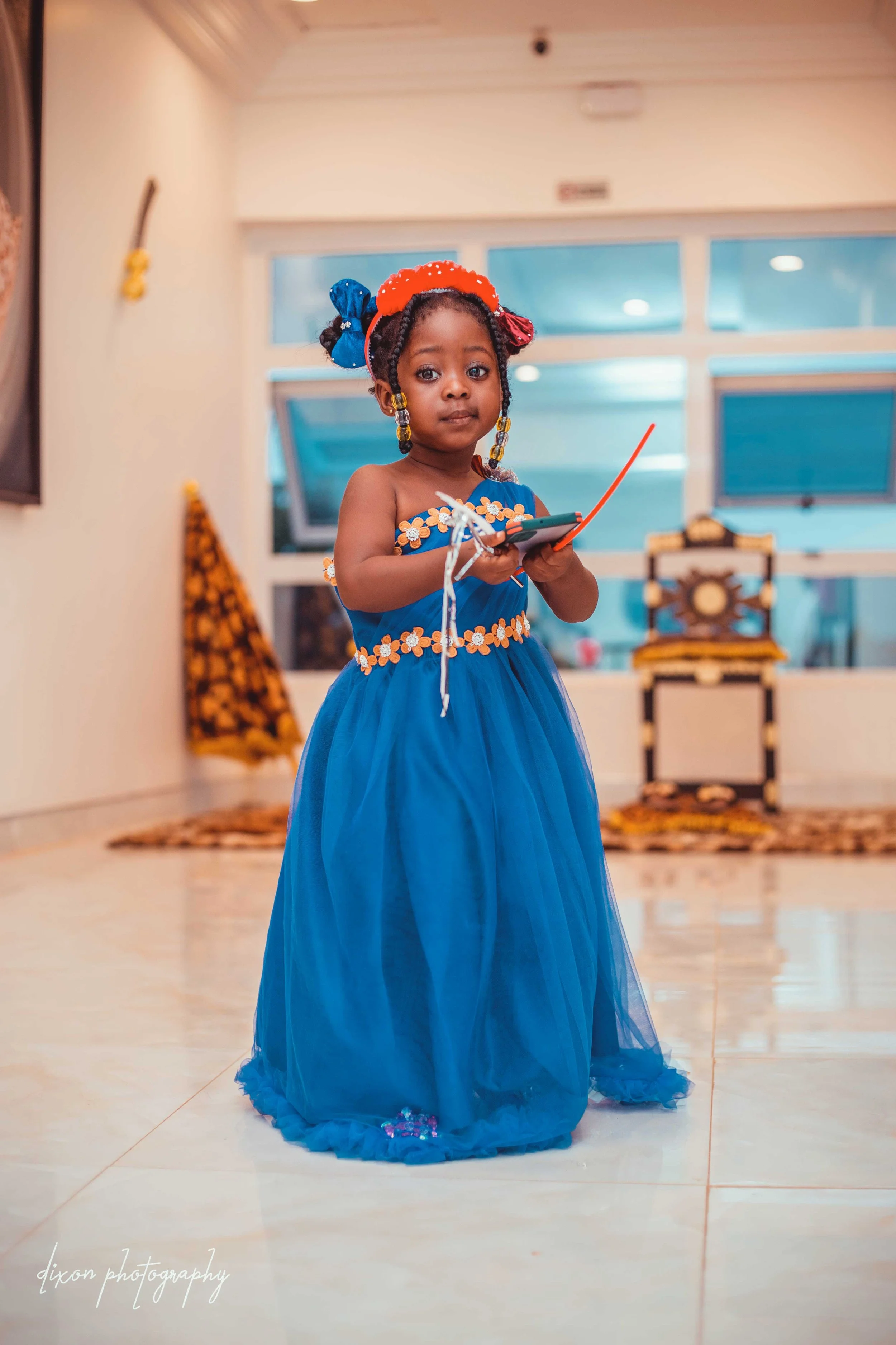 Young girl dressed in a blue princess gown with floral accents, wearing colorful hair accessories, standing indoors while holding a smartphone and a red stick.