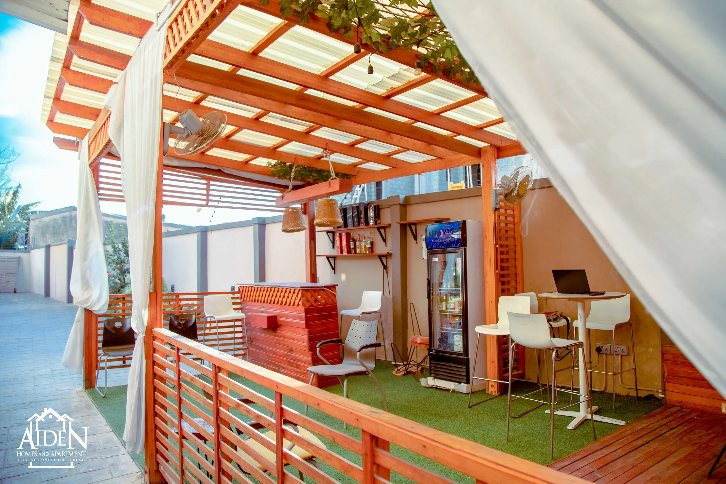 Outdoor bar area with wooden pergola, bar counter, bar stools, high tables, and a refrigerator, surrounded by a privacy fence.