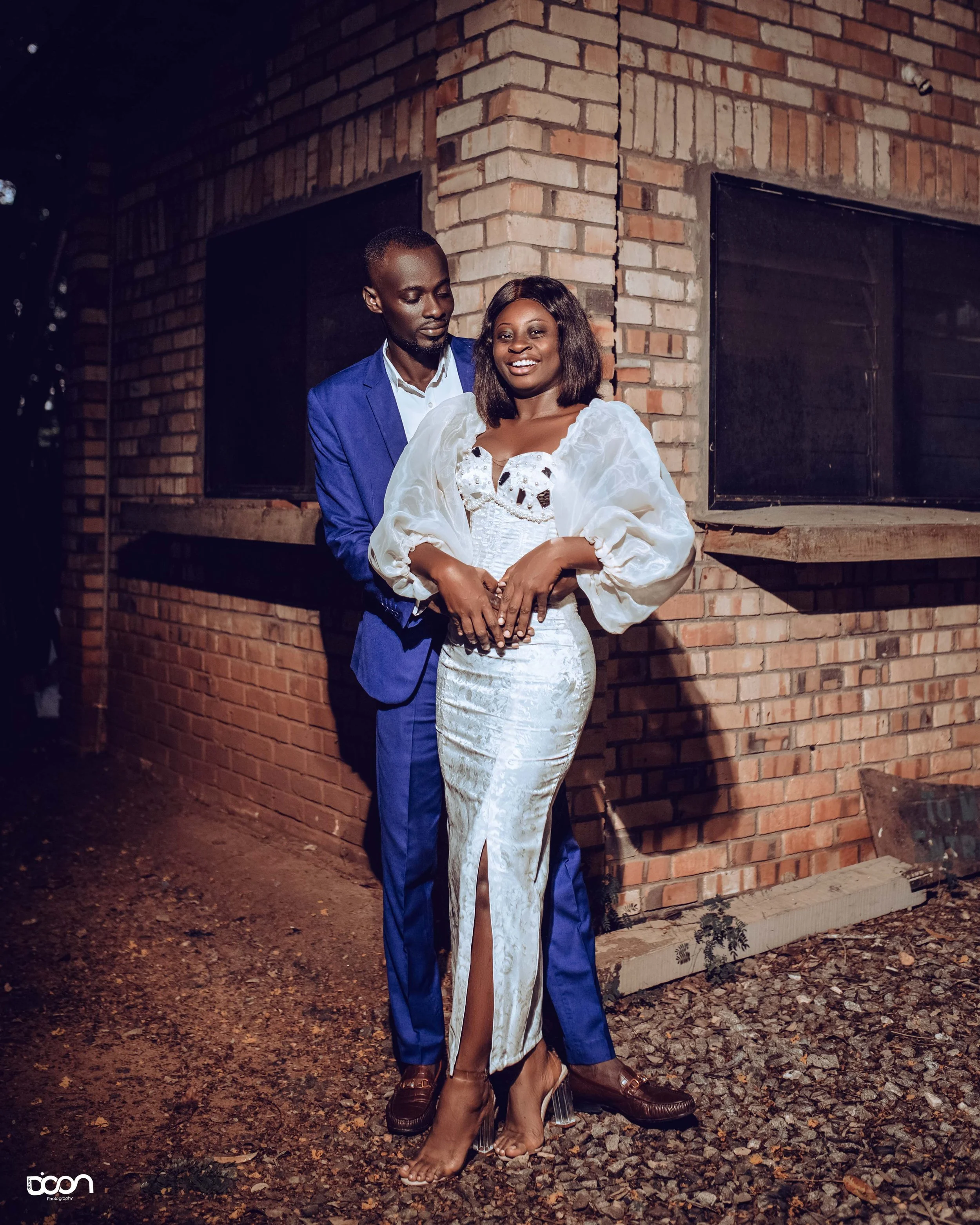 A man and woman standing close together outside at night, smiling, with the man dressed in a blue suit and the woman in a white dress with puffy sleeves, in front of a brick building.