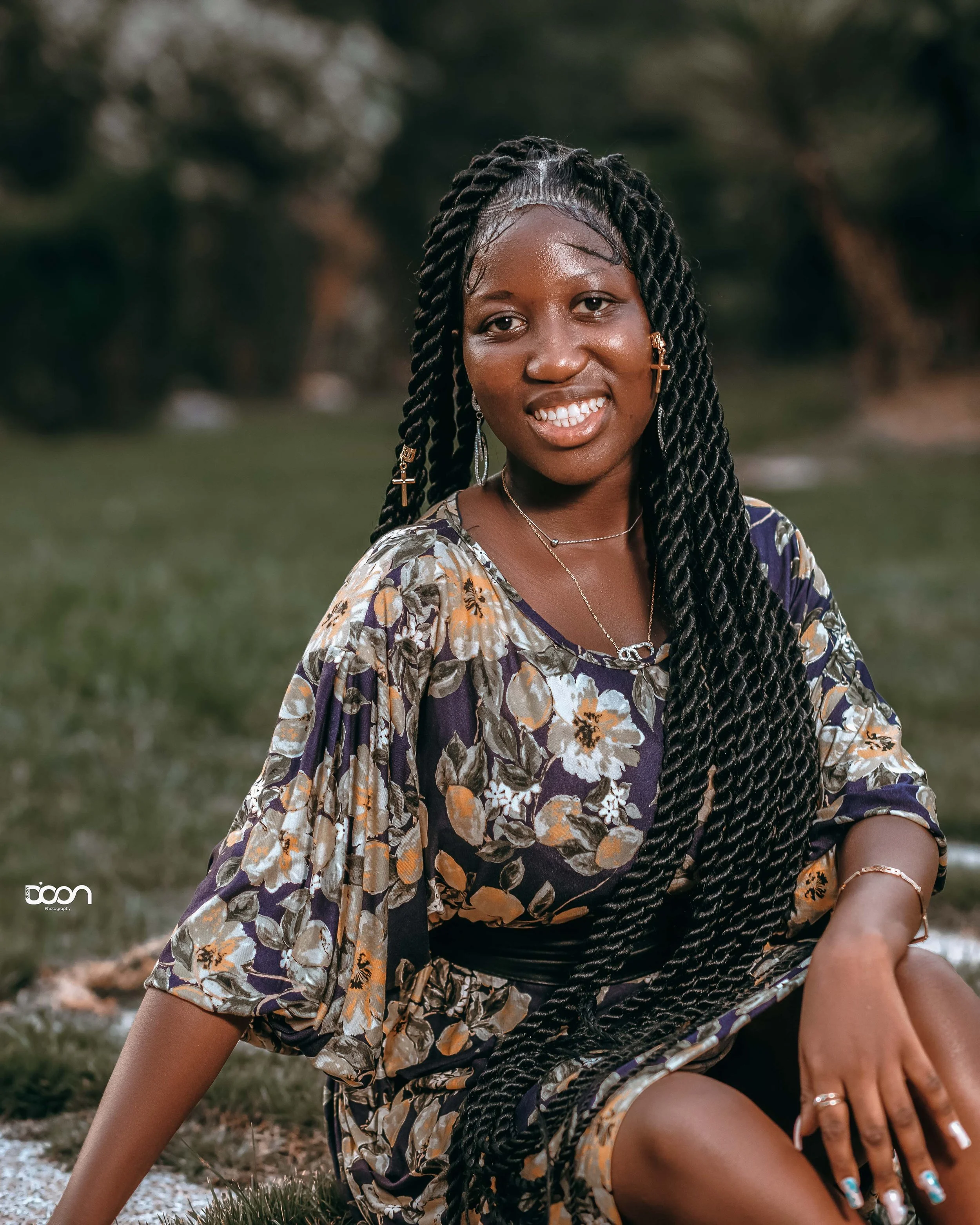 A young woman with long braided hair outdoors, smiling, wearing a floral dress with gold jewelry and cross earrings.