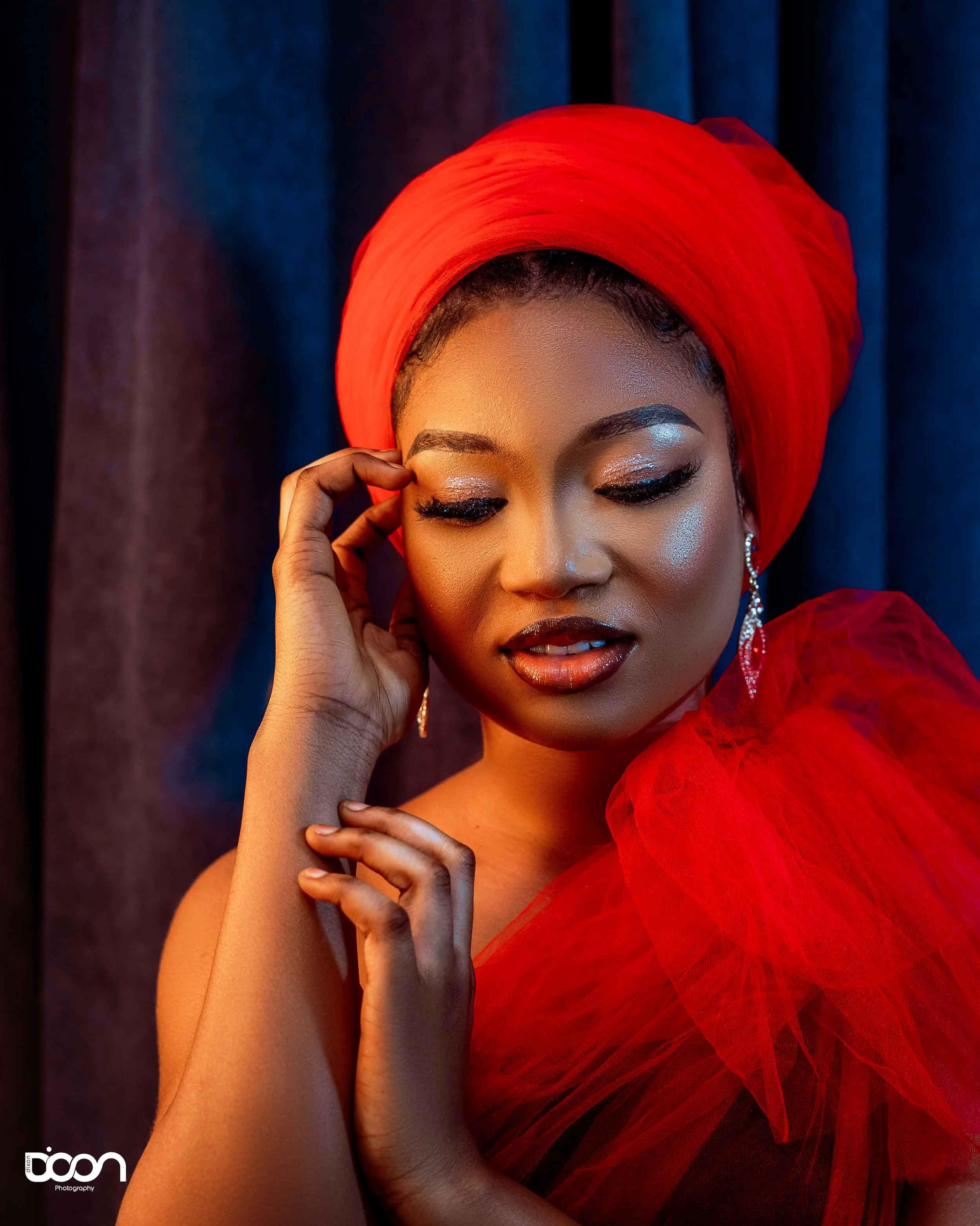 A woman wearing a vibrant red headwrap and matching red tulle dress, with makeup and jewelry, posing with eyes closed in front of dark curtains.