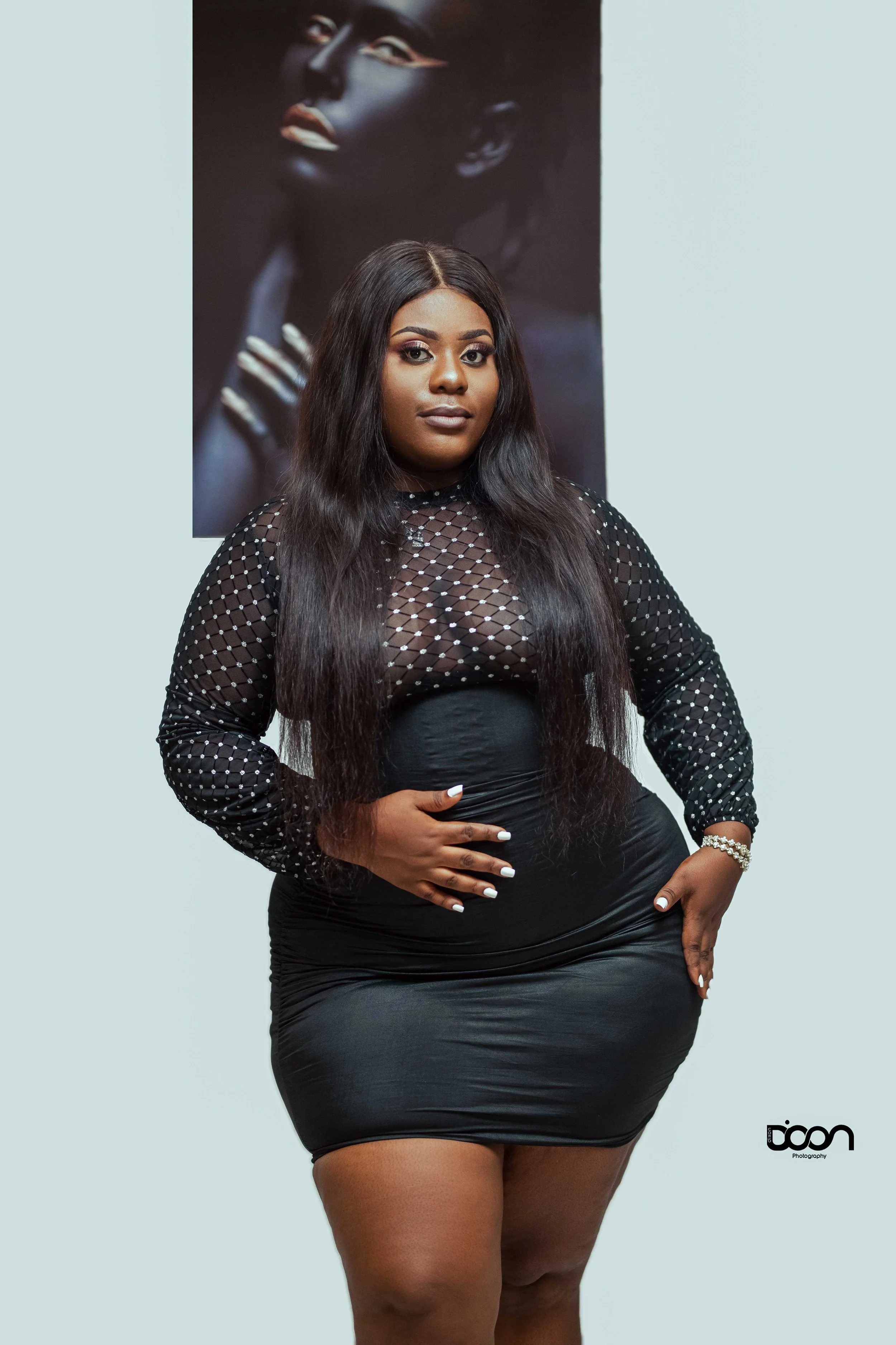 A woman with long black hair wearing a black, semi-transparent, polka-dot top and a tight black skirt, standing in front of a portrait of herself, in a photography studio.