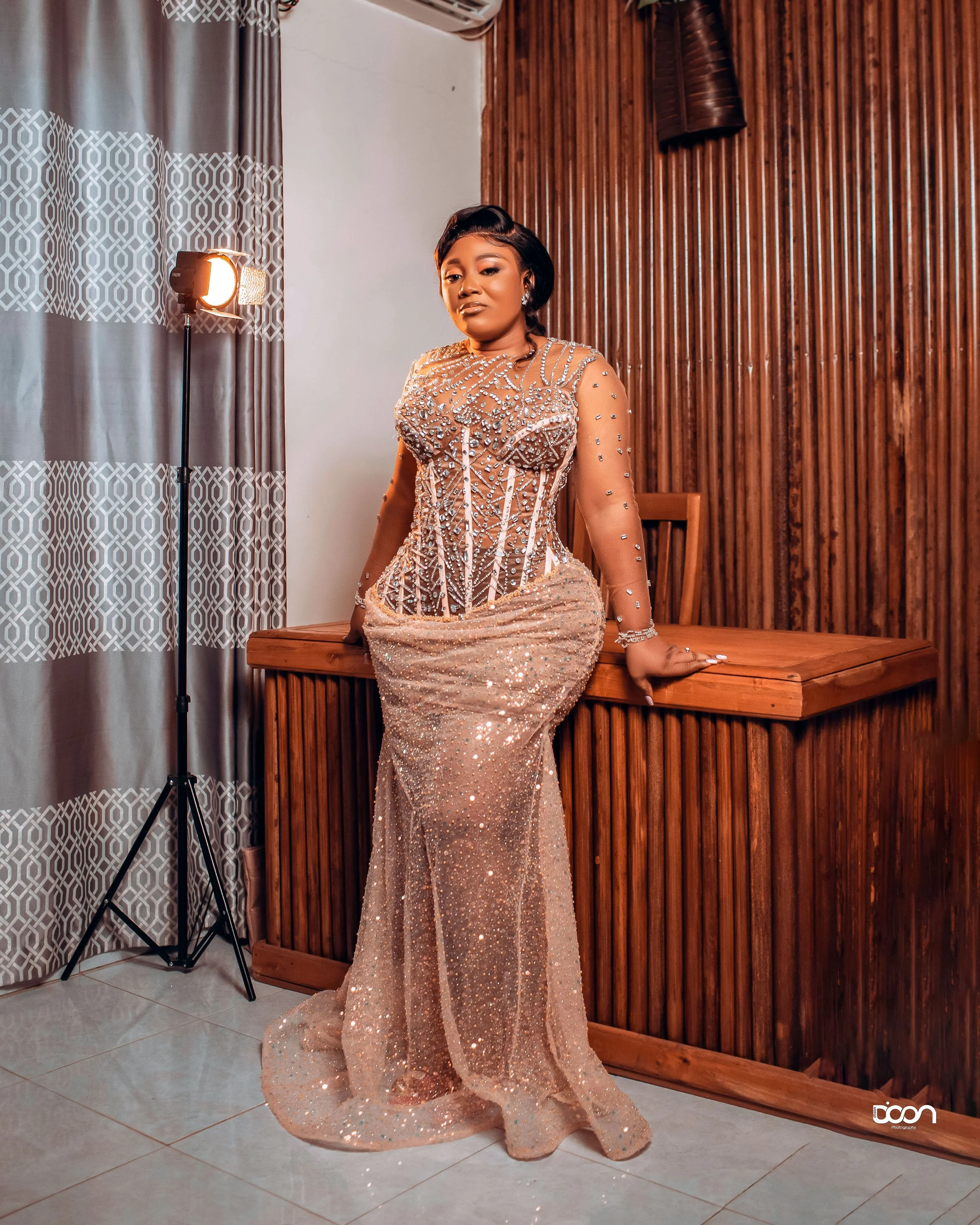 A woman wearing a glamorous, floor-length, gold sequin gown with intricate beadwork, standing in a room with wooden paneling and patterned curtains, posed with one hand resting on a wooden counter.