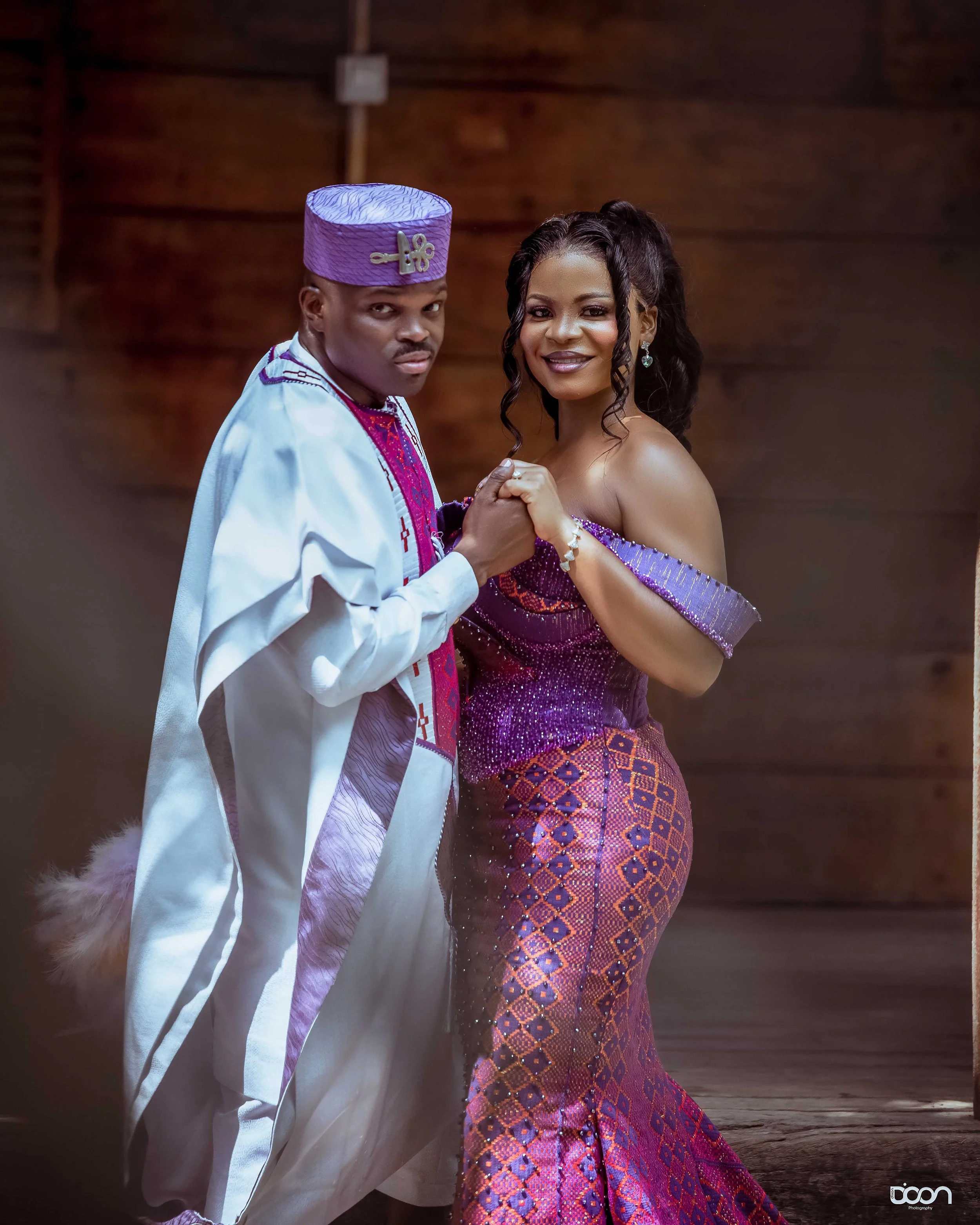 A couple dance together, dressed in traditional African attire, in front of a wooden background.