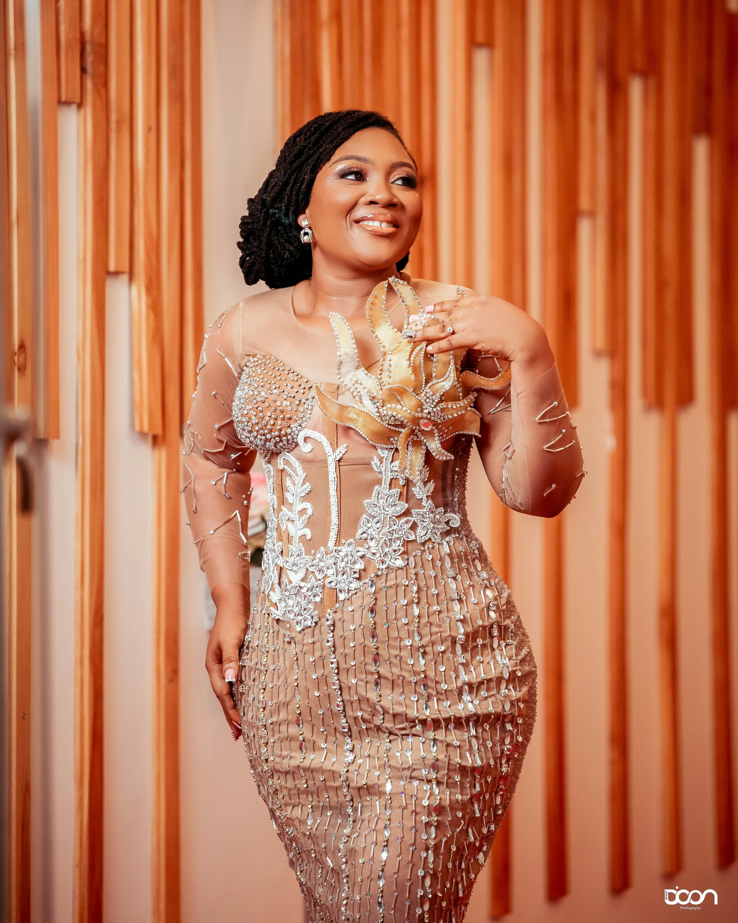 A woman dressed in a beige, embellished dress with intricate beadwork and floral details, standing indoors near a wooden wall, smiling and touching her shoulder.
