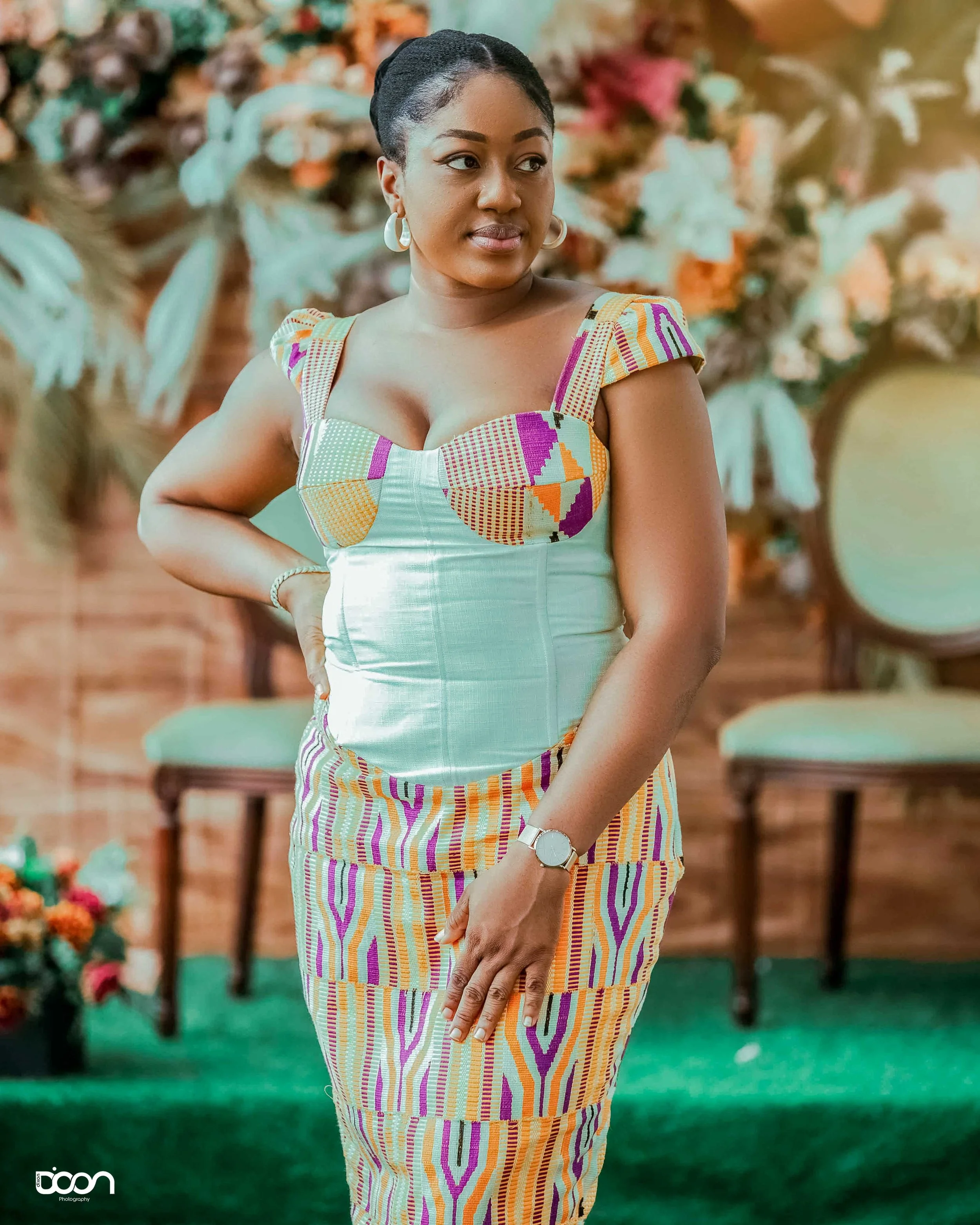 A woman wearing a colorful, patterned dress with a fitted bodice and puffy sleeves, standing in front of a floral backdrop and chairs, posing with one hand on her hip and looking to the side.