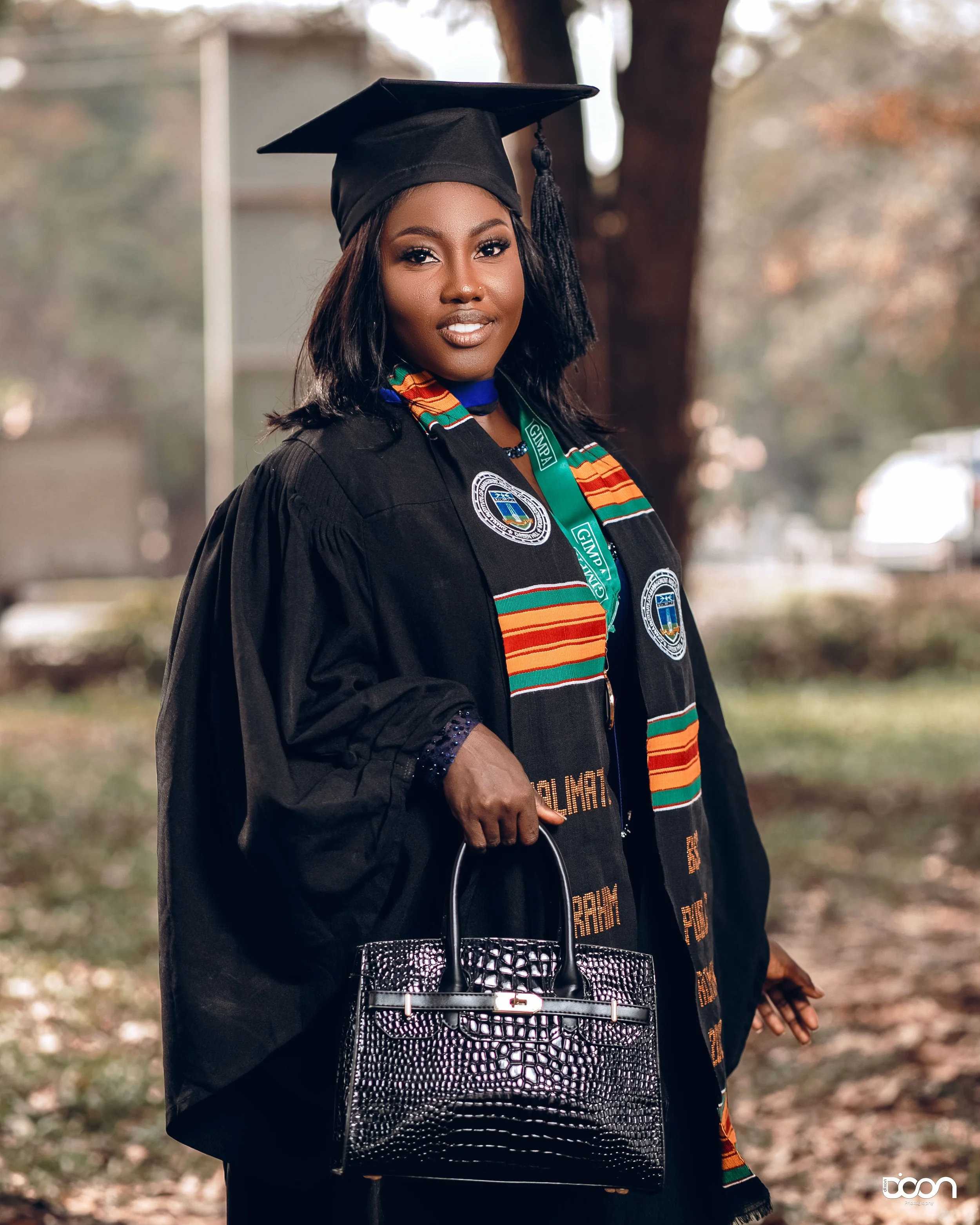 A woman in a graduation cap and gown holding a black handbag, outdoors with trees and a blurred background.
