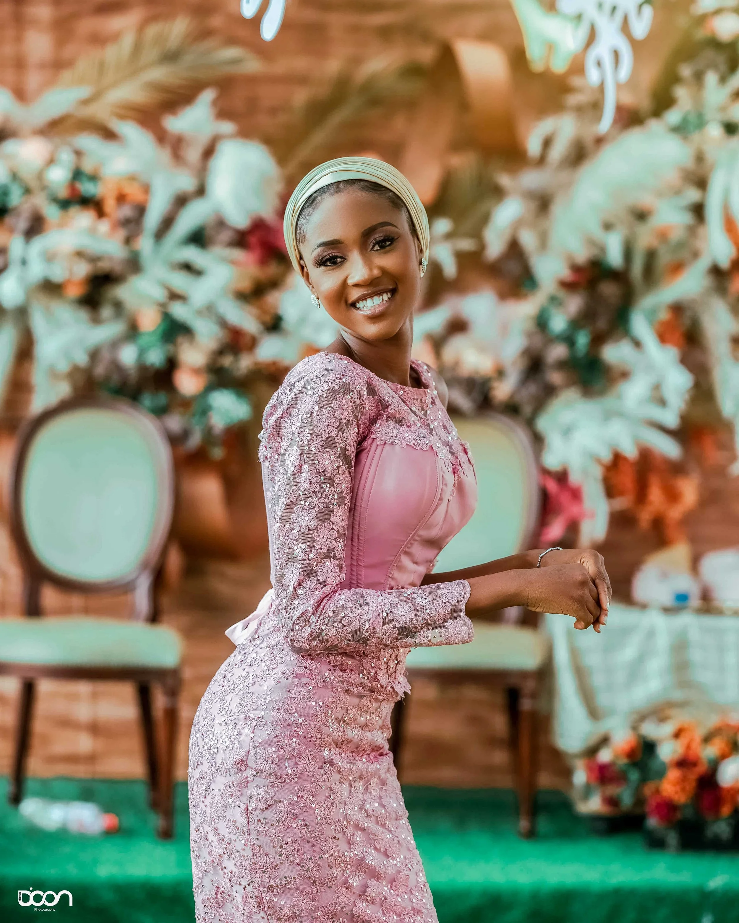 A woman smiling at a celebration, wearing a pink lace dress and a headwrap, standing in front of a floral and wooden backdrop with chairs and decorations.