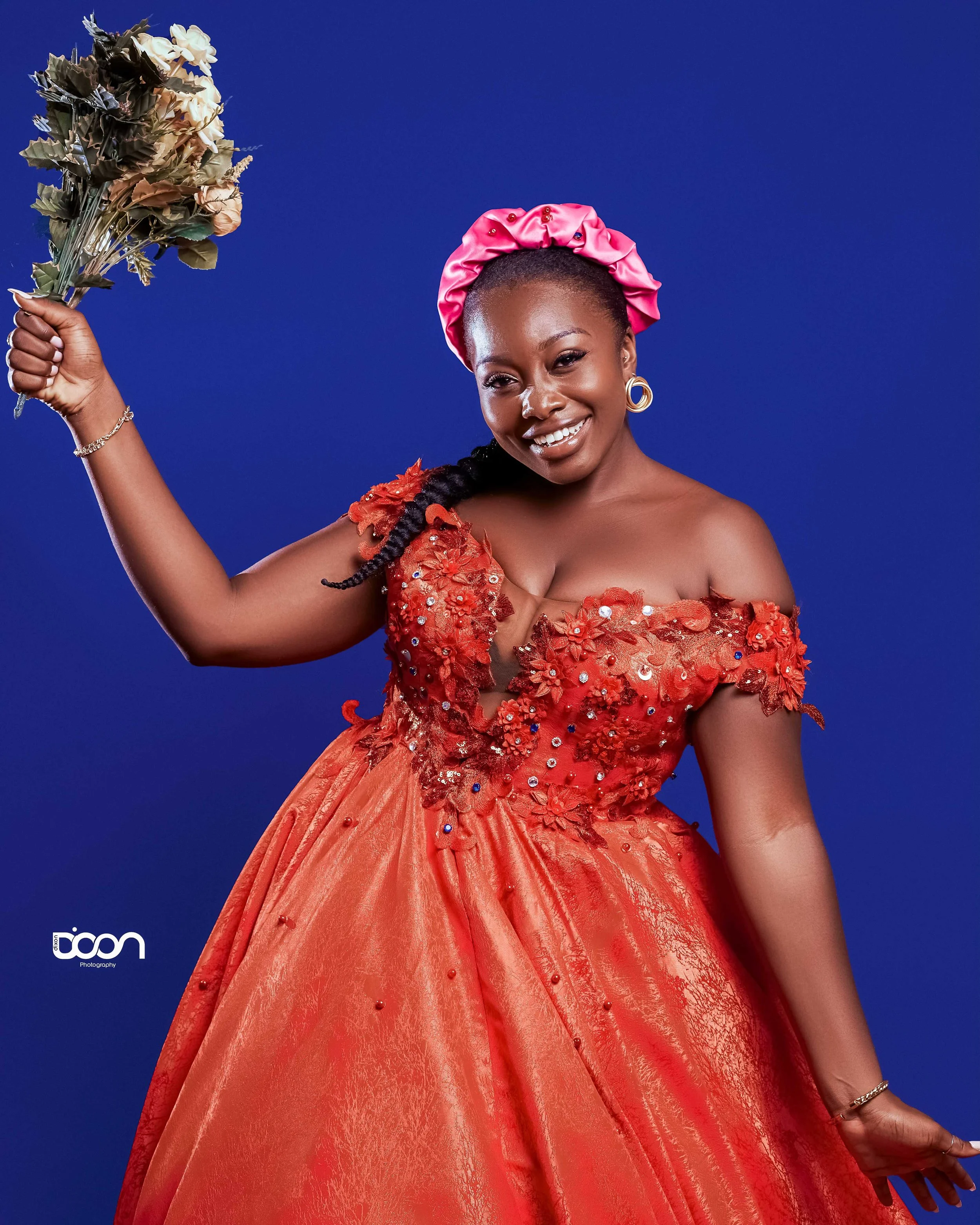 A woman wearing a red, floral, off-the-shoulder gown with embellishments, pink satin headwrap, smiling, holding a bouquet of dried flowers, against a blue background.