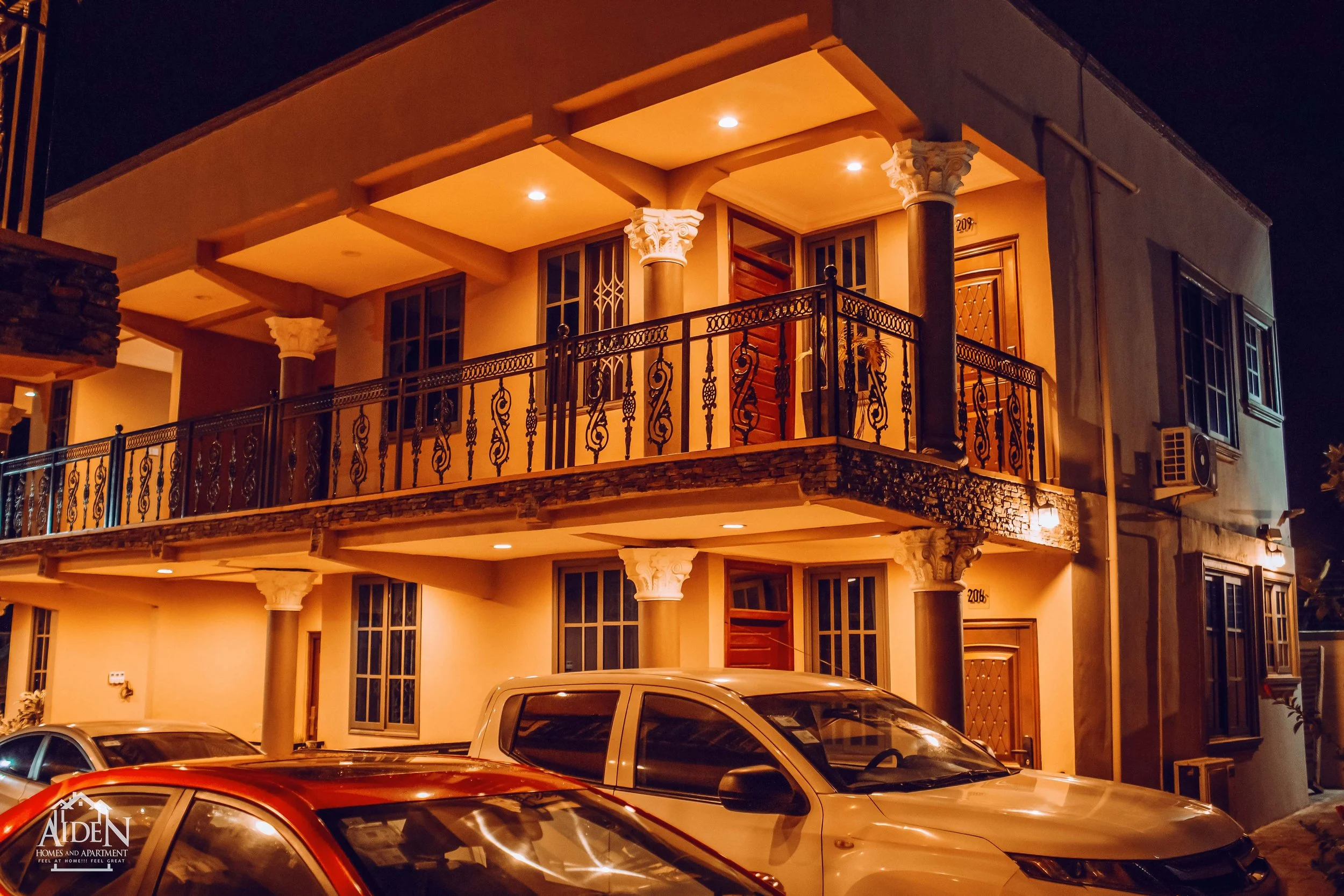 Two-story apartment building at night with warm lighting, black decorative balcony railings, and parked cars in front.
