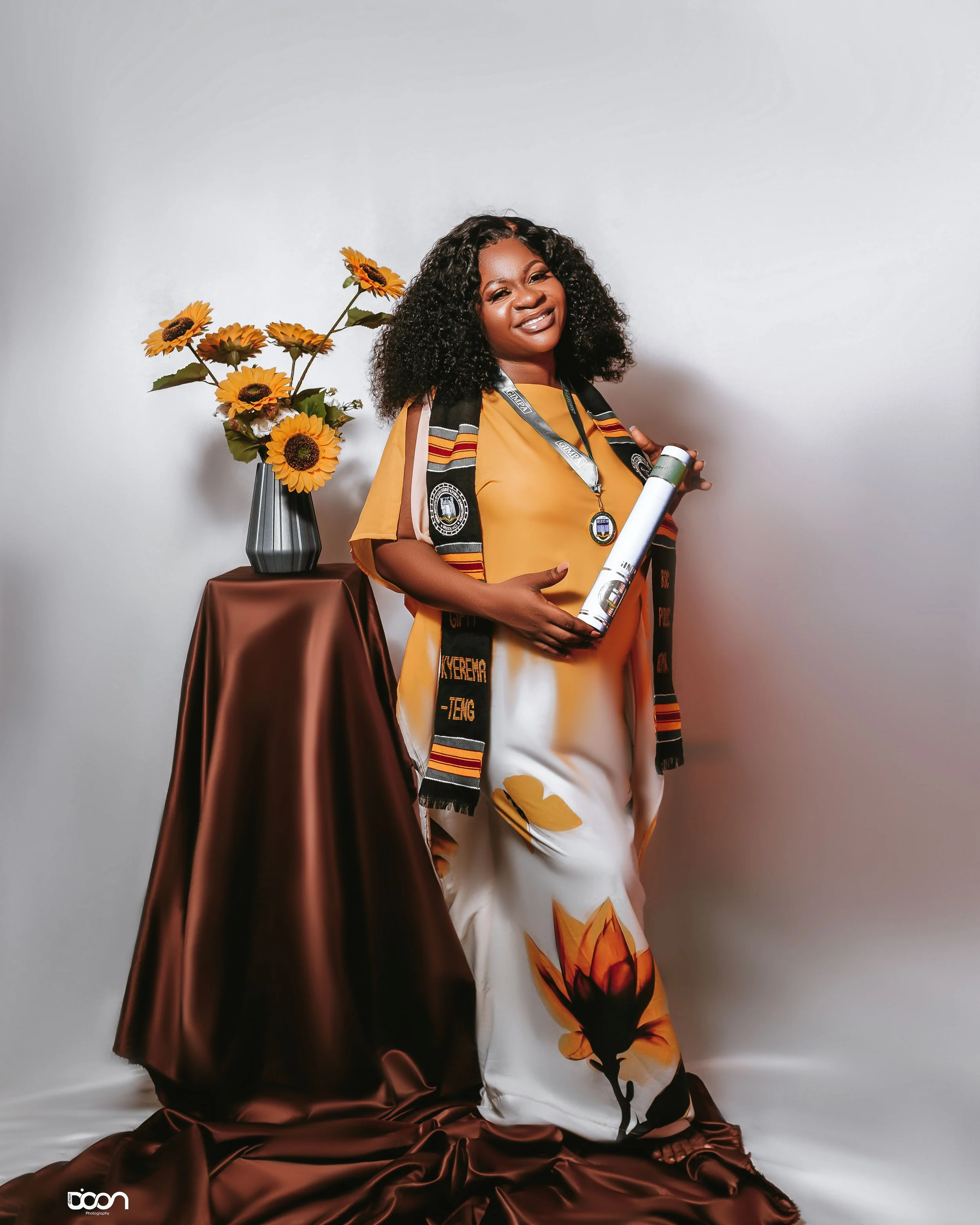 Woman in graduation gown and sash holding diploma, standing next to a table with a vase of sunflowers on a brown satin cloth against a plain white background.