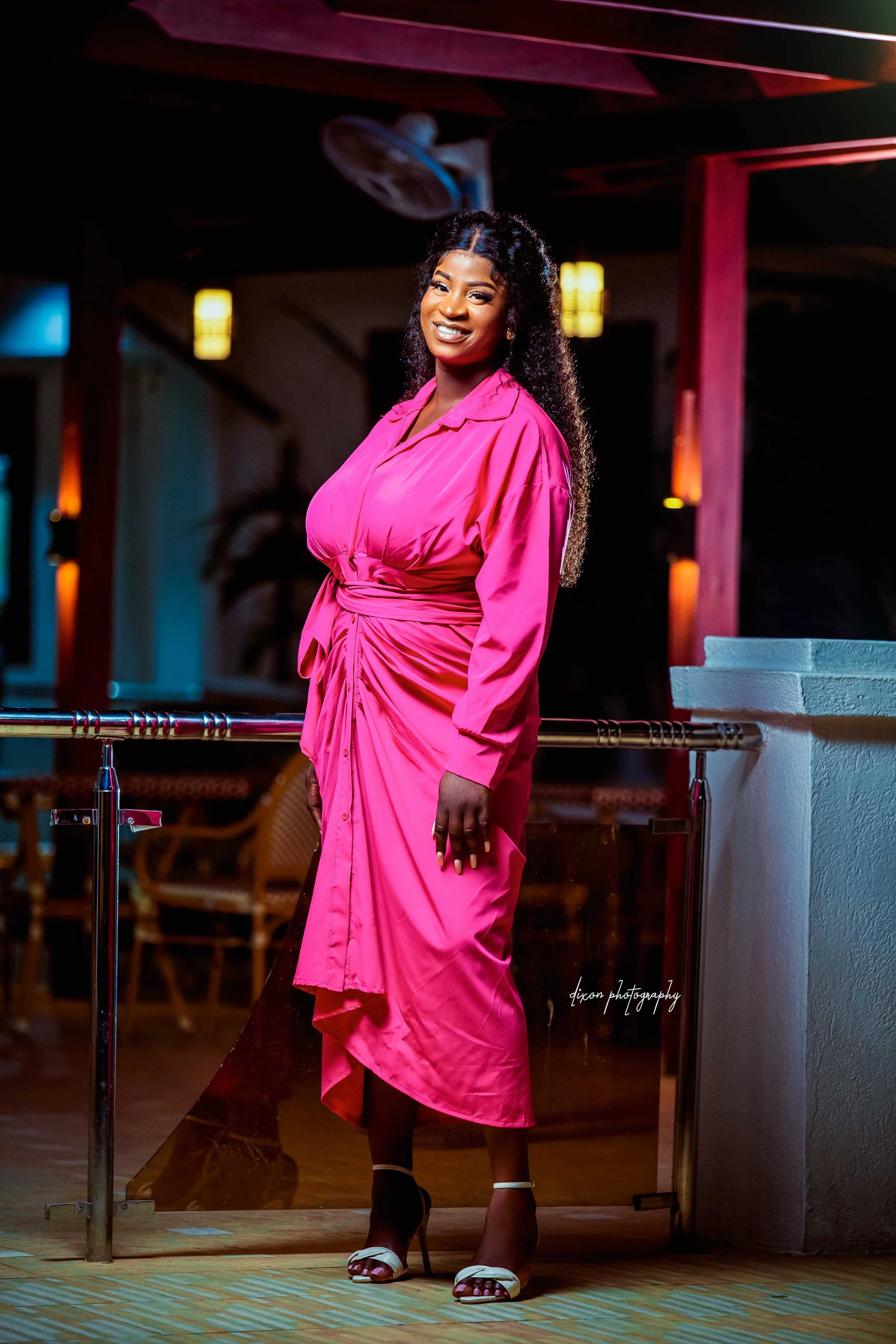 A woman in a pink dress standing indoors with a smile, dark curly hair, high heels, and warm lighting.