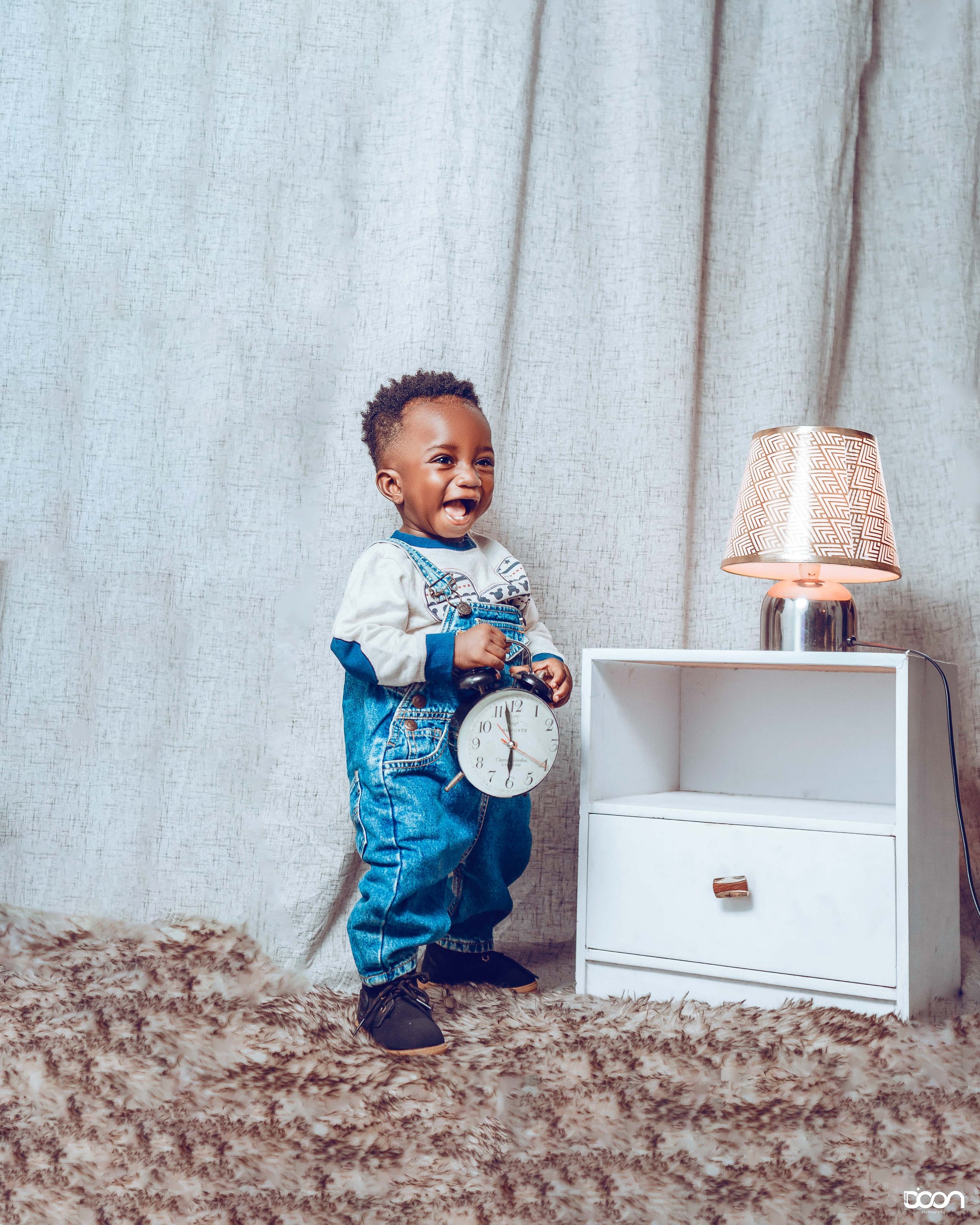 A young boy wears denim overalls over a long-sleeved shirt, holds a large clock, and laughs in a cozy room with a white nightstand and a lamp.