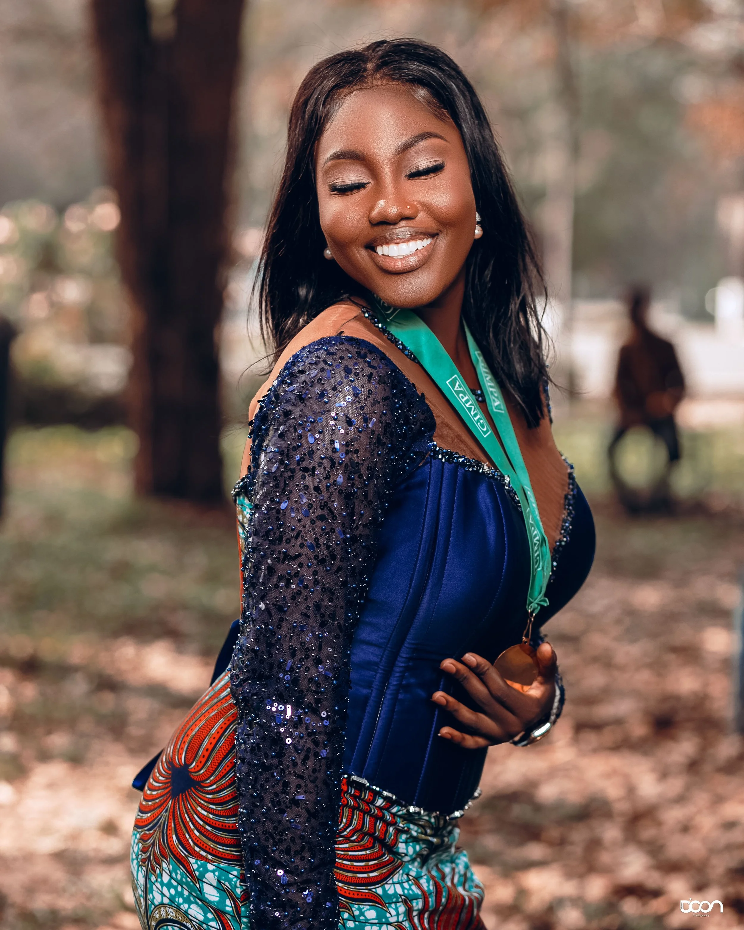 Smiling woman in colorful traditional attire and a blue dress with a green graduation sash outdoors.