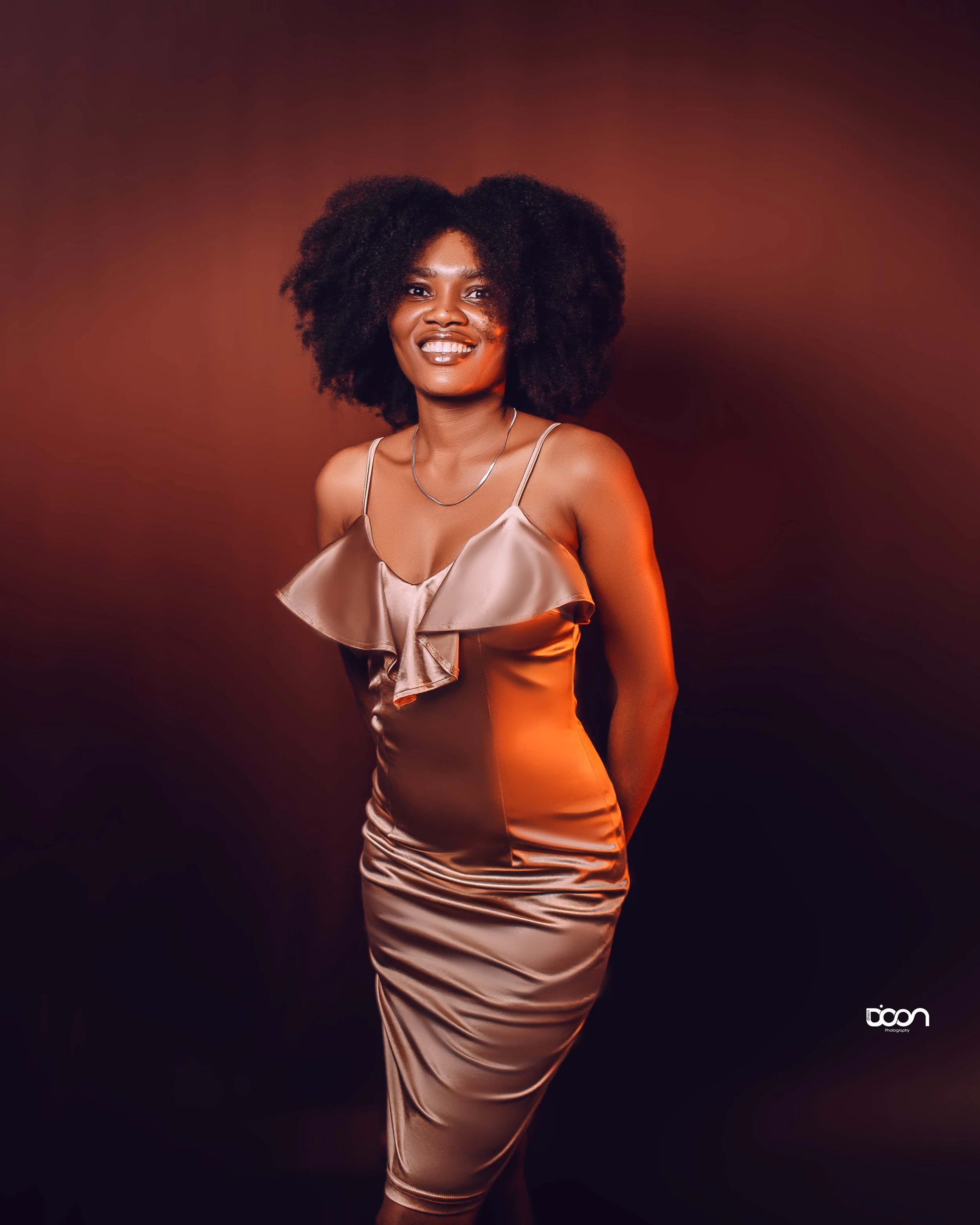 A woman with dark skin and voluminous natural hair, wearing a silky, beige sleeveless dress standing against a dark, gradient background, smiling at the camera.