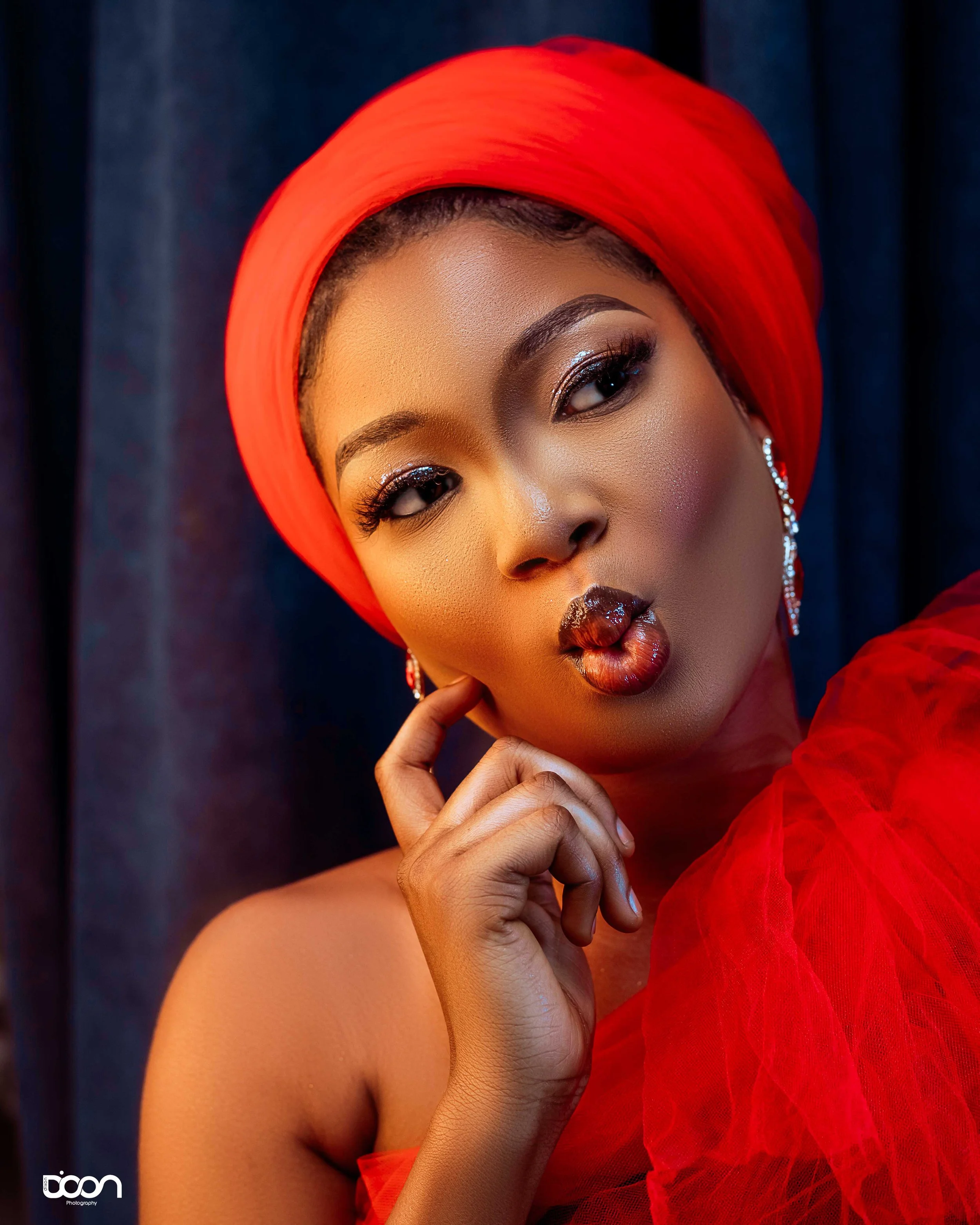 A woman with dark skin wearing a red head wrap and red tulle dress, making a kiss face with shiny dark lipstick, with earrings and makeup.