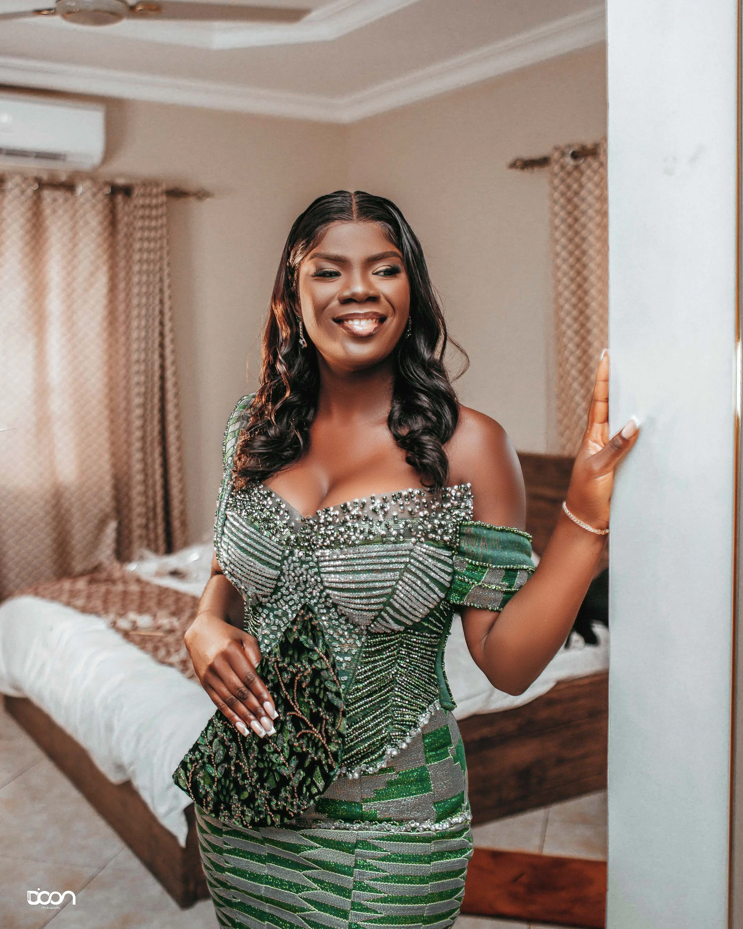 A smiling woman in a decorated green and silver dress standing in a bedroom, holding a mirror.