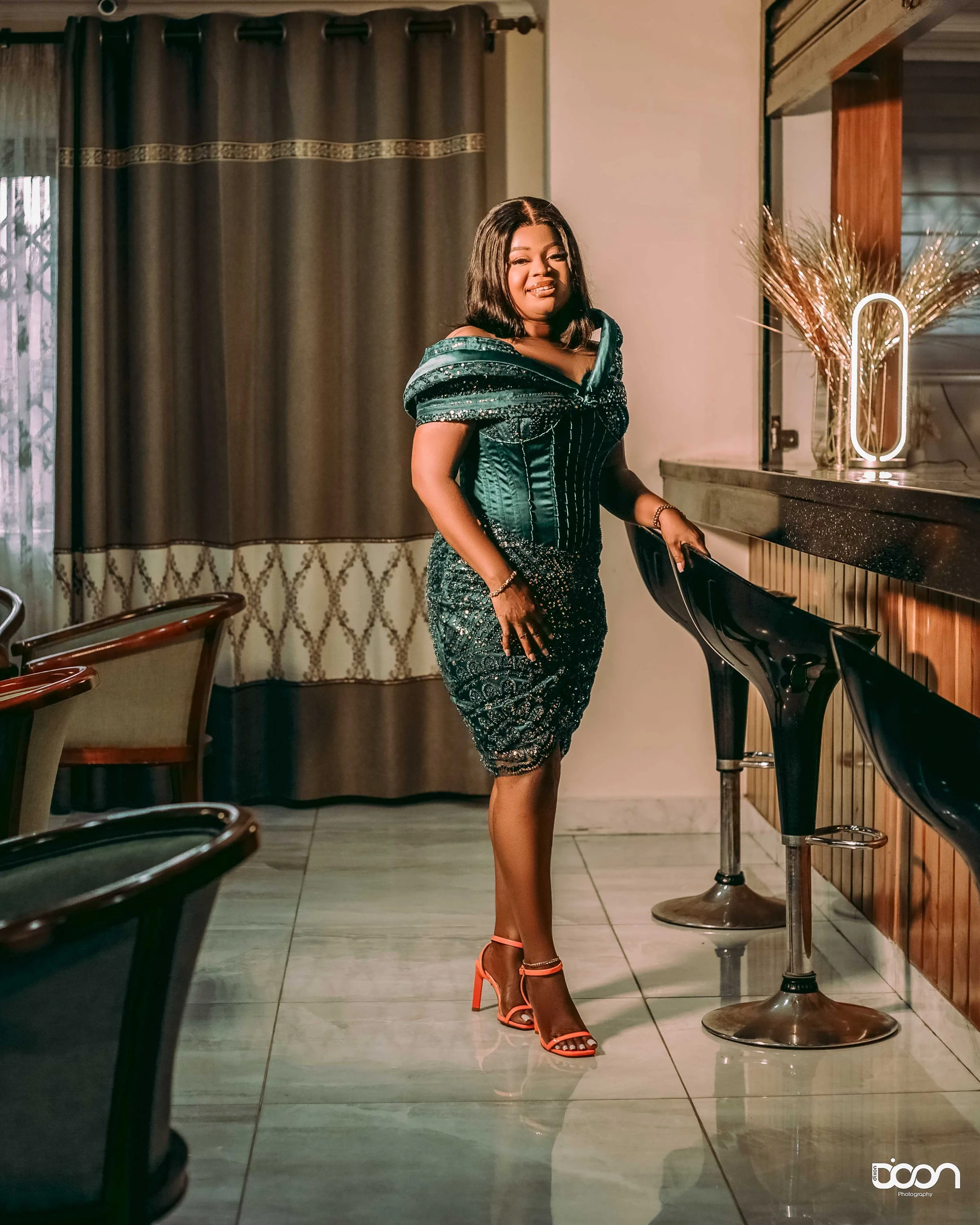 A woman in an elegant dark green and black dress with orange high-heeled shoes standing next to a bar counter in a well-lit room.