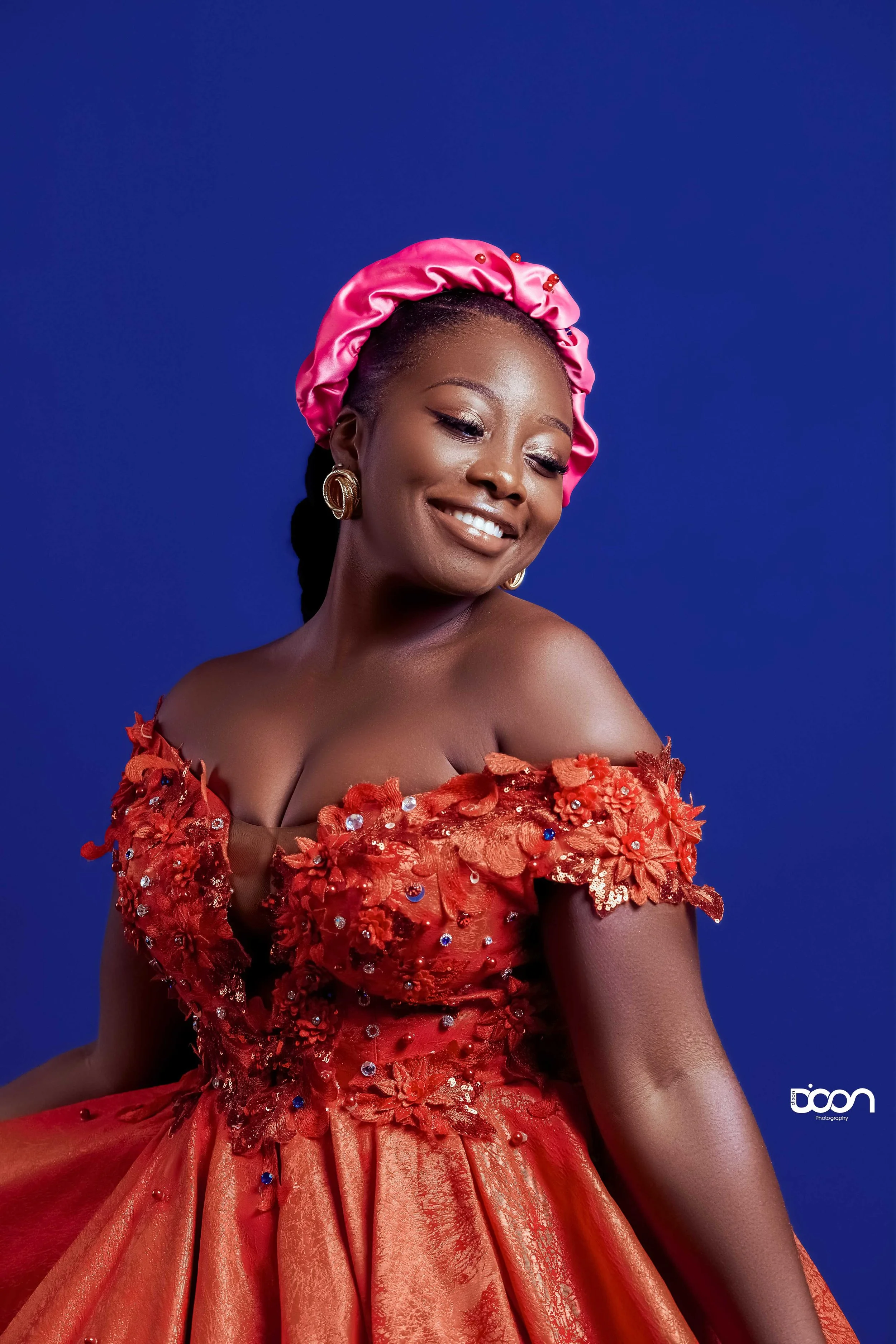 A woman smiling, wearing a red, off-shoulder gown with floral and bead embellishments, accessorized with gold earrings and a pink satin bonnet, against a solid blue background.