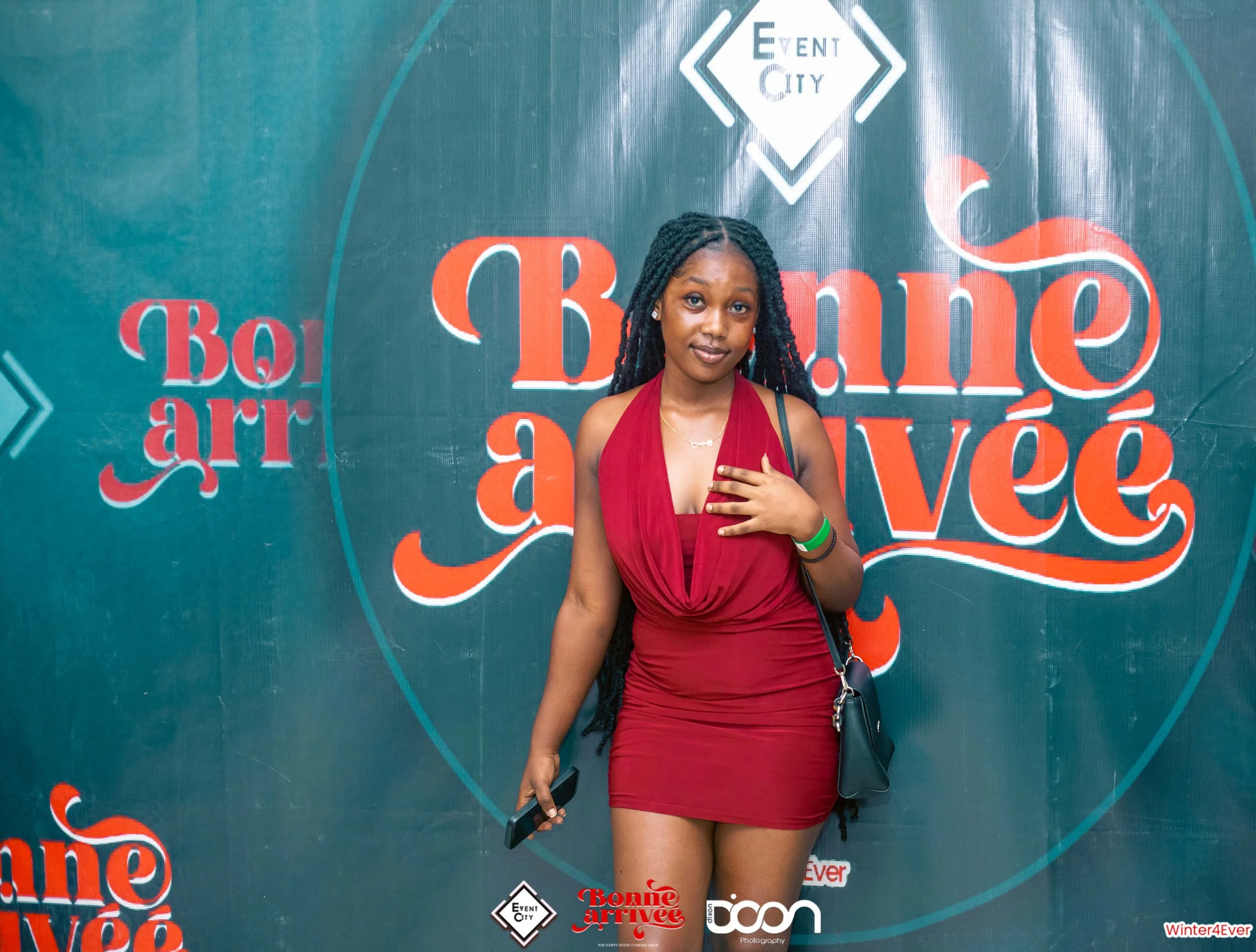 Young woman in red dress standing in front of a sign that reads 'Bonne arrivée' at a themed event, holding a phone and wearing a green wristband.