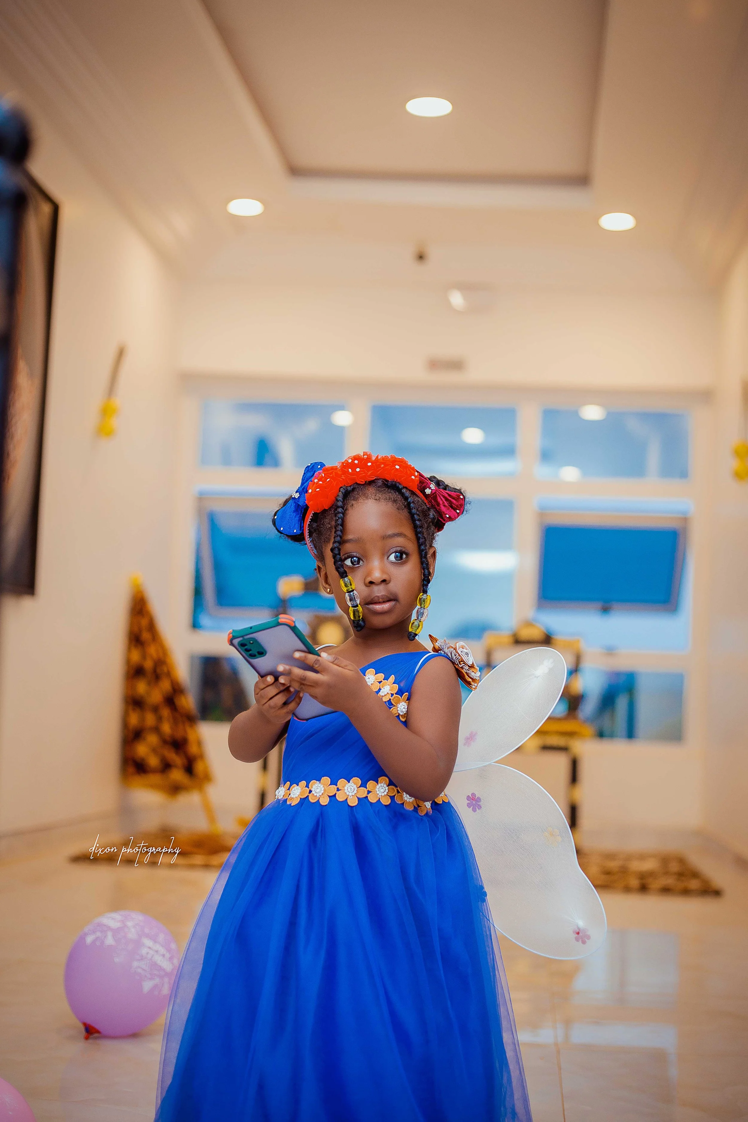 A young girl dressed as a fairy, wearing a blue dress with flower embellishments, fairy wings, and a colorful headband, holding a phone indoors with balloons on the floor.