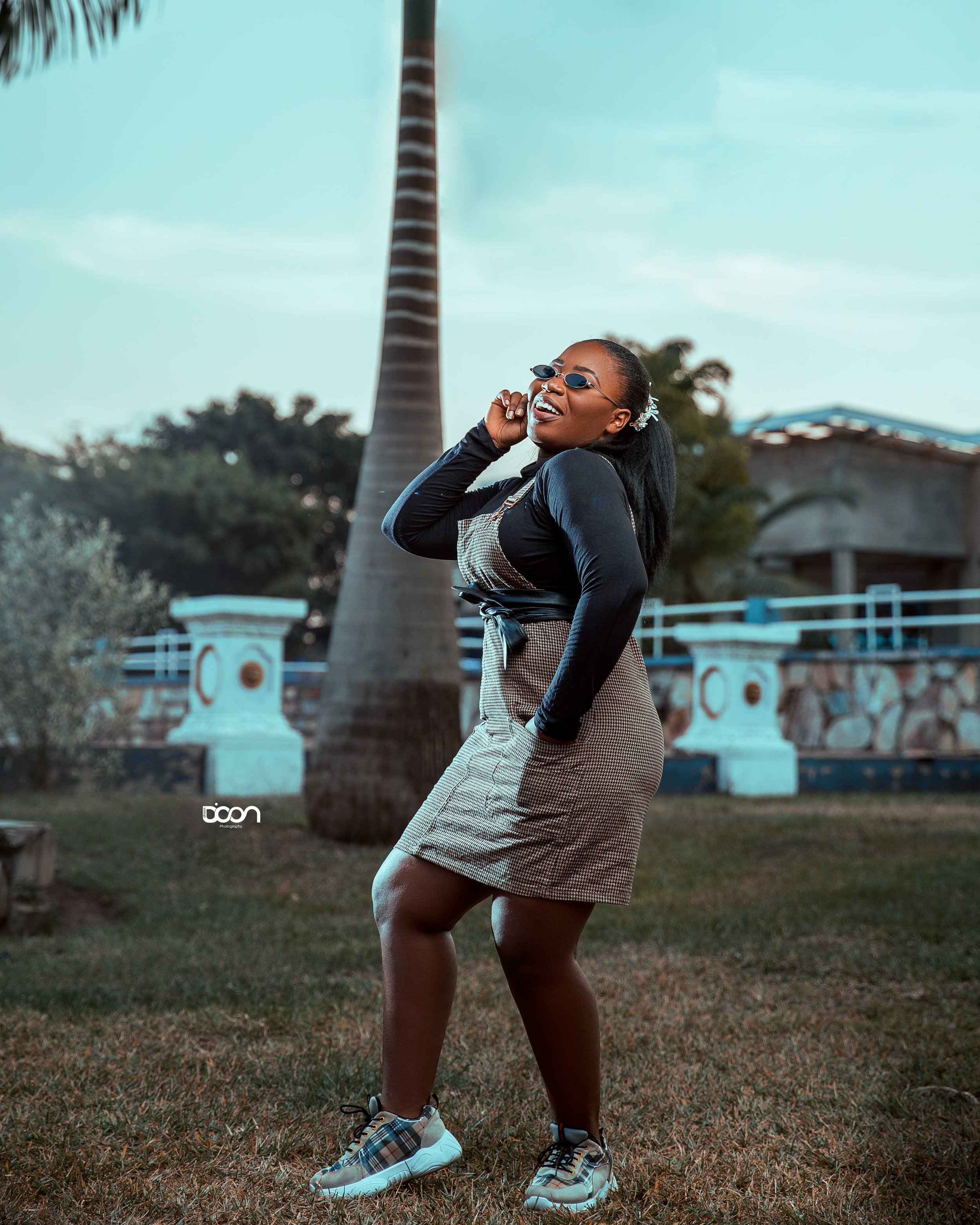 A woman is standing outdoors on grass, smiling and talking on her phone. She is wearing sunglasses, a black long-sleeve top, a checked skirt, and sneakers. There are palm trees and a small building in the background.