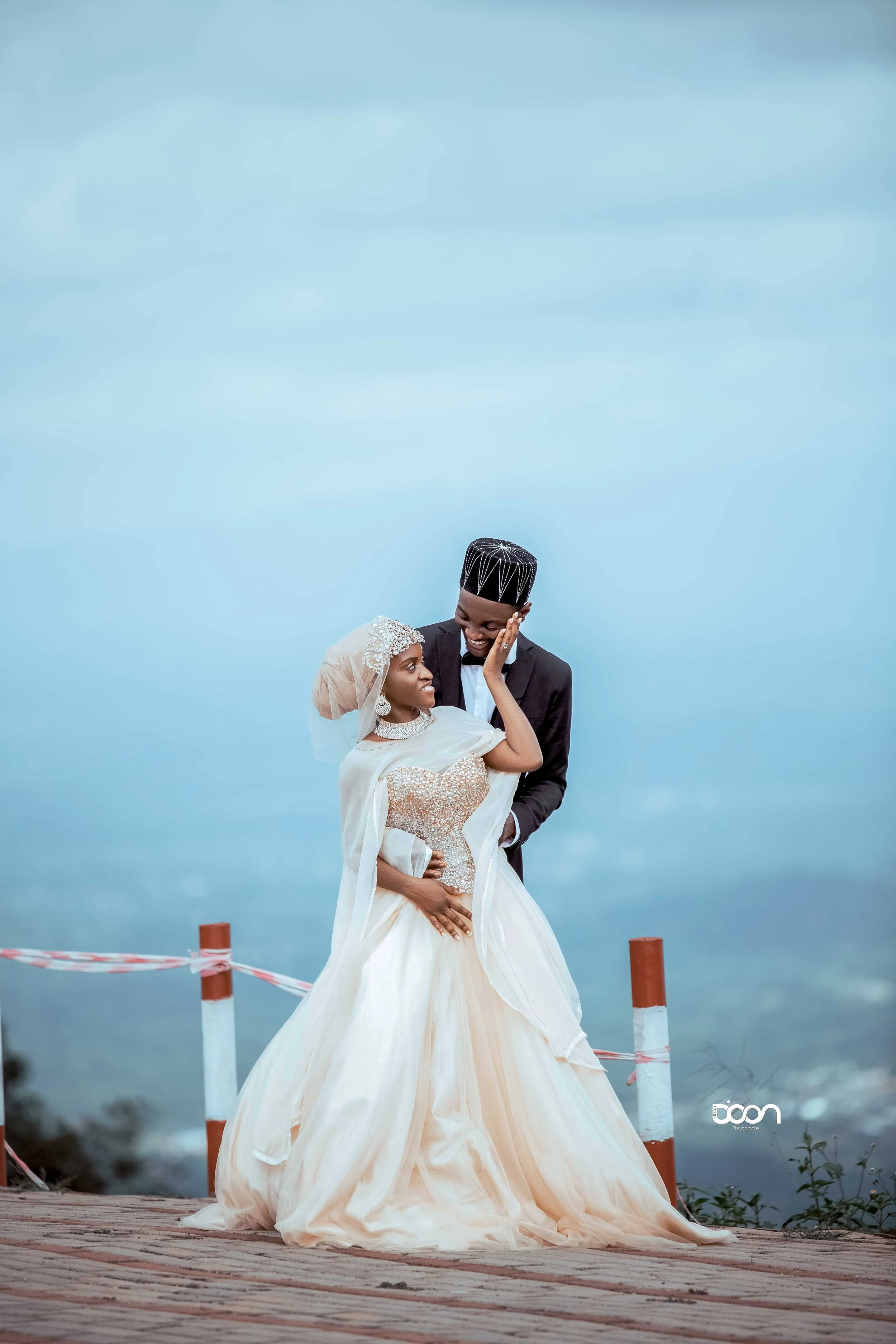 A bride and groom in traditional wedding attire smiling and sharing a tender moment outdoors on a cloudy day with a scenic view in the background.