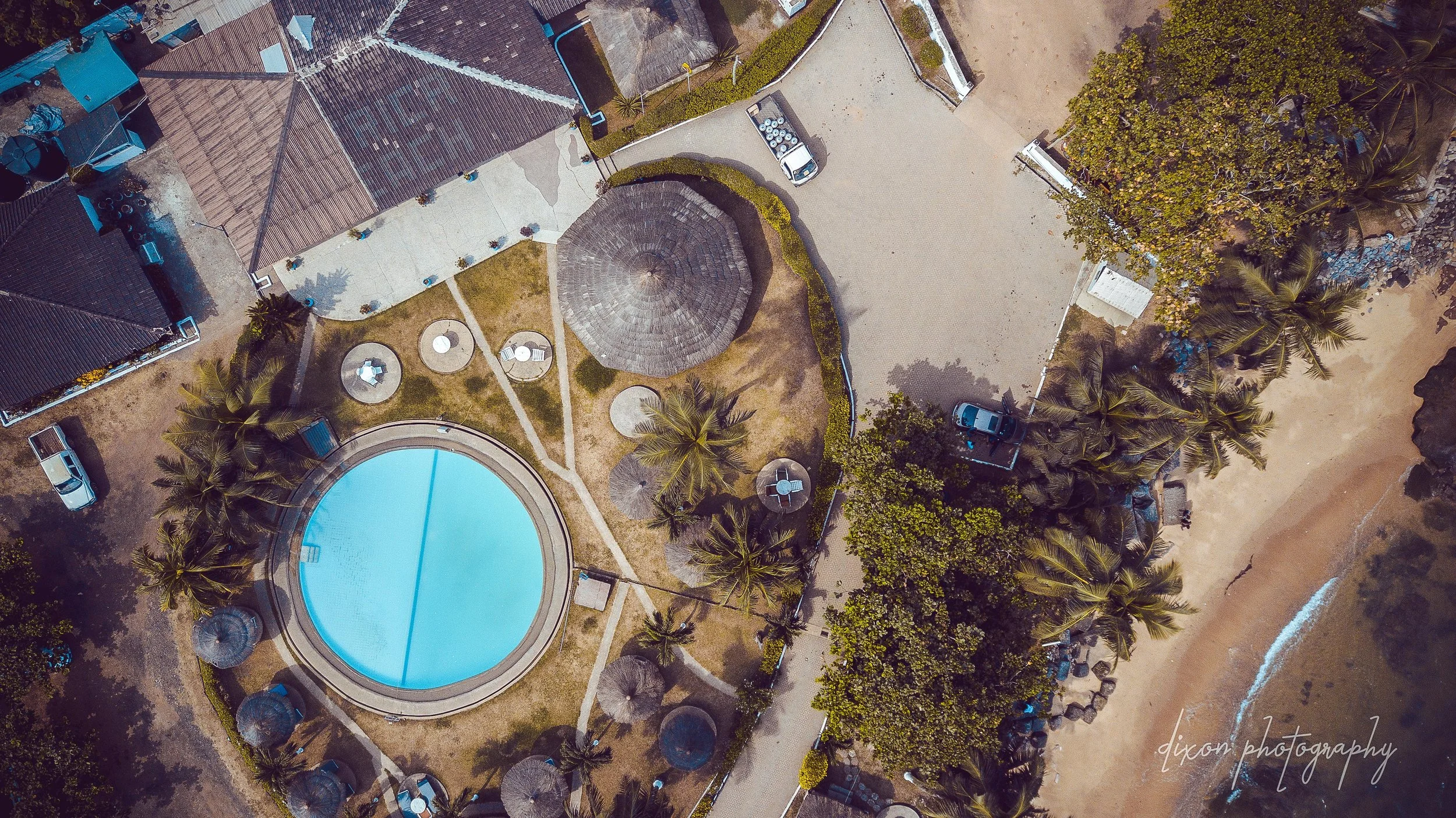 An aerial view of a resort area with a large round swimming pool surrounded by palm trees, straw umbrellas, and lounge chairs, next to a sandy beach with a few cars parked nearby.