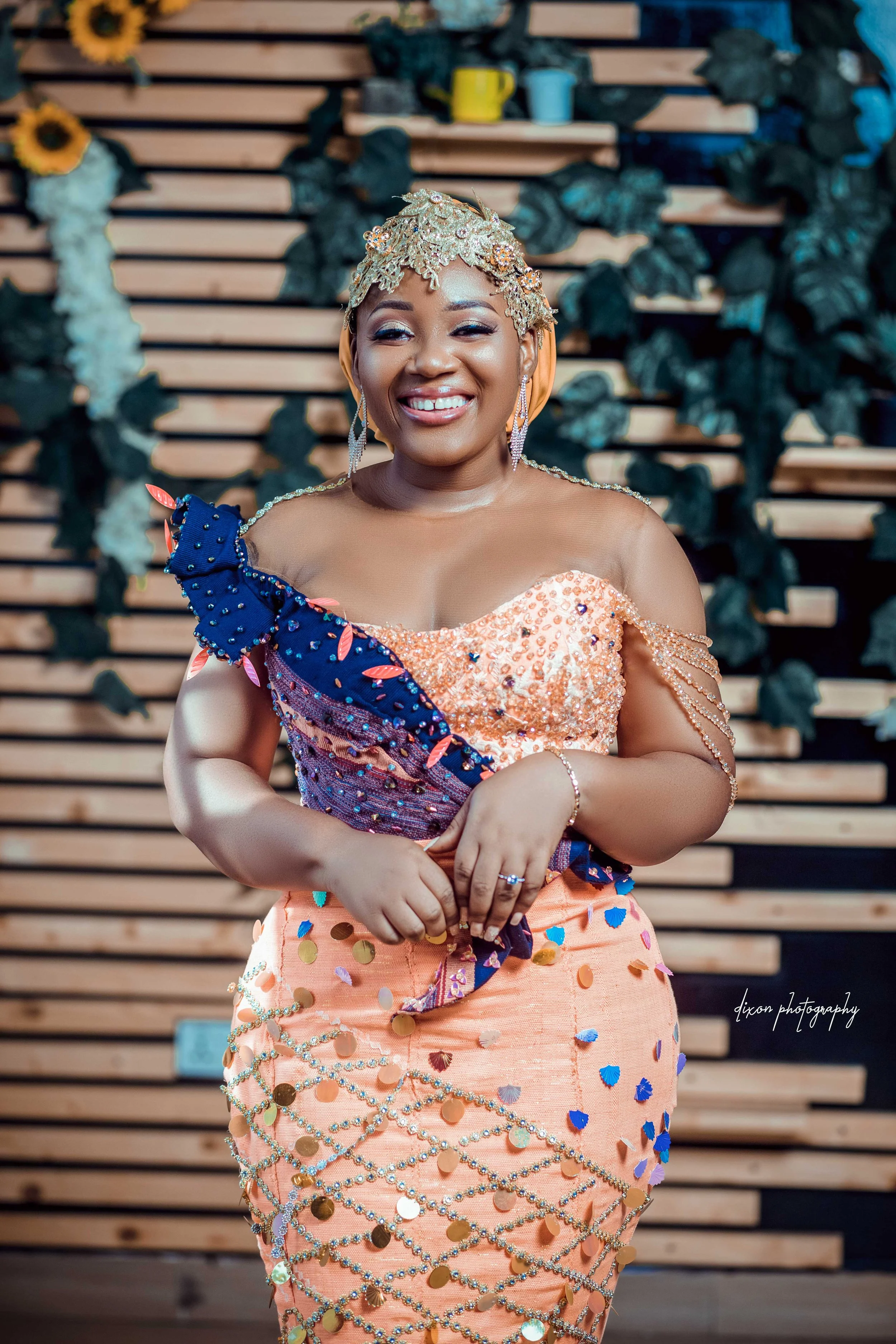 A woman smiling wearing a peach-colored dress decorated with sequins and beads, with a beaded chain on the skirt, and a blue and pink sash over her shoulder. She has a gold floral headpiece and earrings, and stands in front of a wooden wall with gree