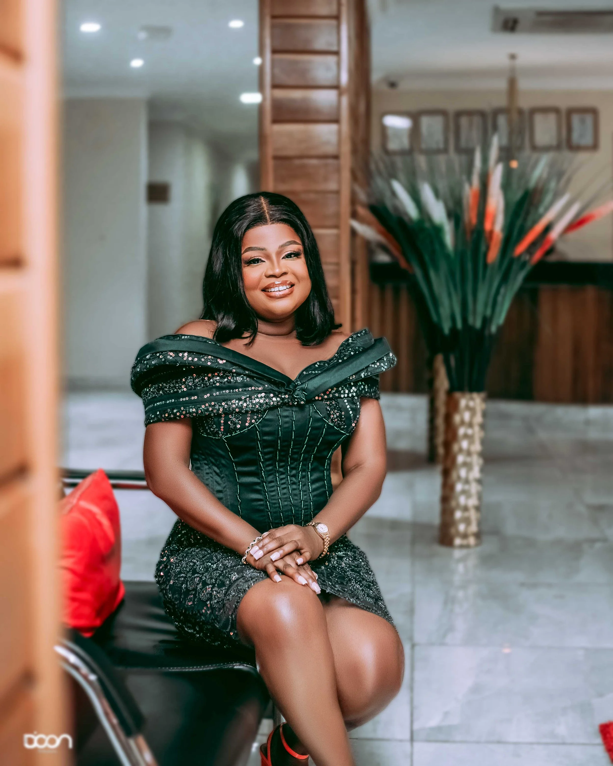 A woman in a black, sparkly off-the-shoulder dress sits on a black chair, smiling at the camera indoors with wooden wall decor and a large floral arrangement in the background.