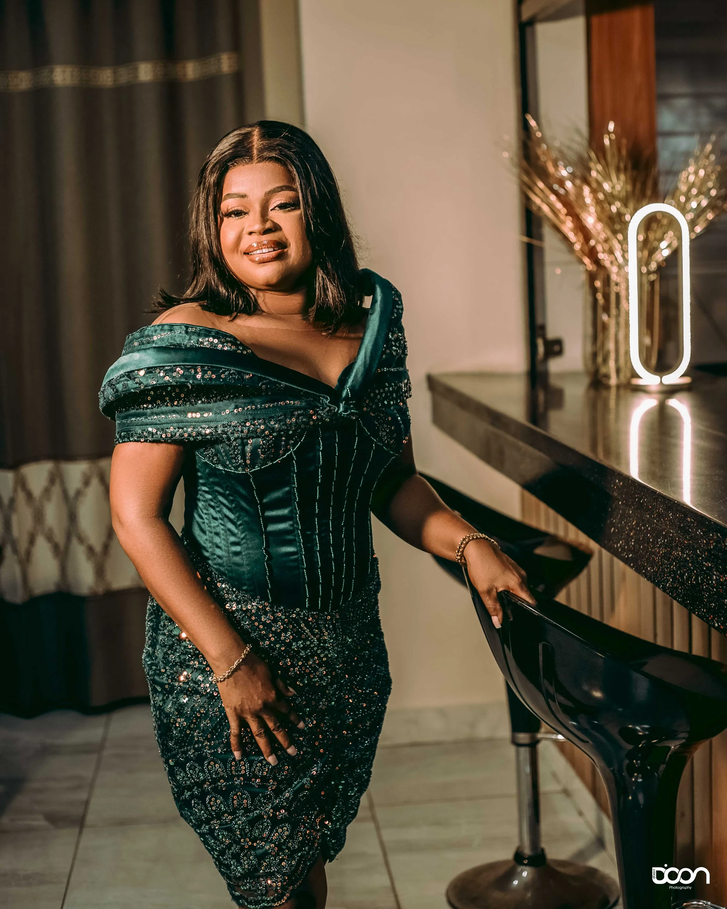 A woman in an elegant, dark green, sparkly dress with off-the-shoulder design, standing beside a black bar stool in a warmly lit room with decorative art pieces and a mirror with a modern oval-shaped light.