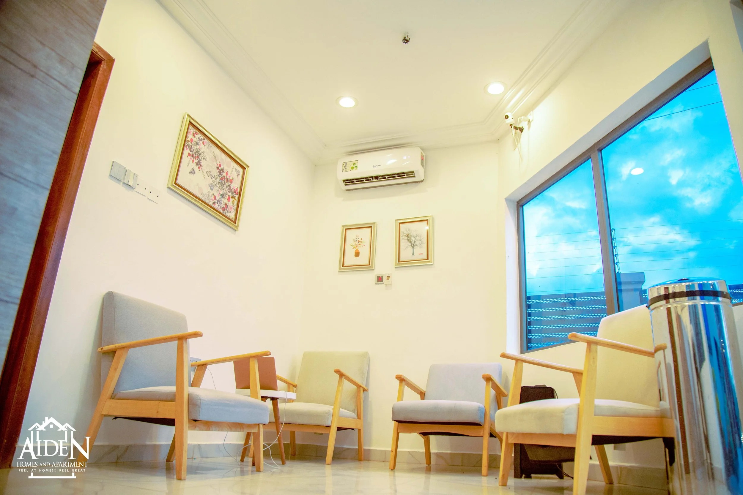 A waiting room with four wooden-framed chairs with light-colored cushions, a small table with a laptop and a white cable, framed artwork on the wall, an air conditioning unit, a large window showing a blue sky with clouds, and a shiny metal trash bin